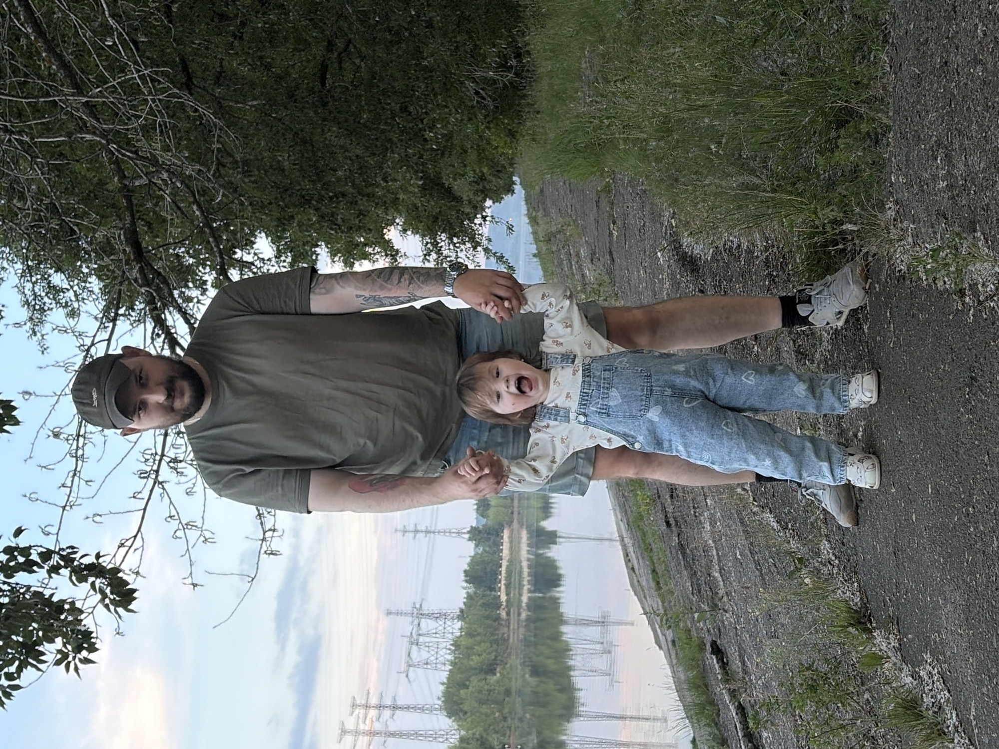 An adult male wearing a green t-shirt, denim shorts, and a baseball cap stands on a paved path next to a body of water. He is holding the hands of a young child who is wearing blue denim overalls and a white long-sleeved shirt. The child is facing the camera with their mouth open and arms outstretched. The background shows a tree-lined shore, a calm body of water, and several power lines in the distance. The location is the embankment of the Kama River in the city of Chaikovsky, Russia.