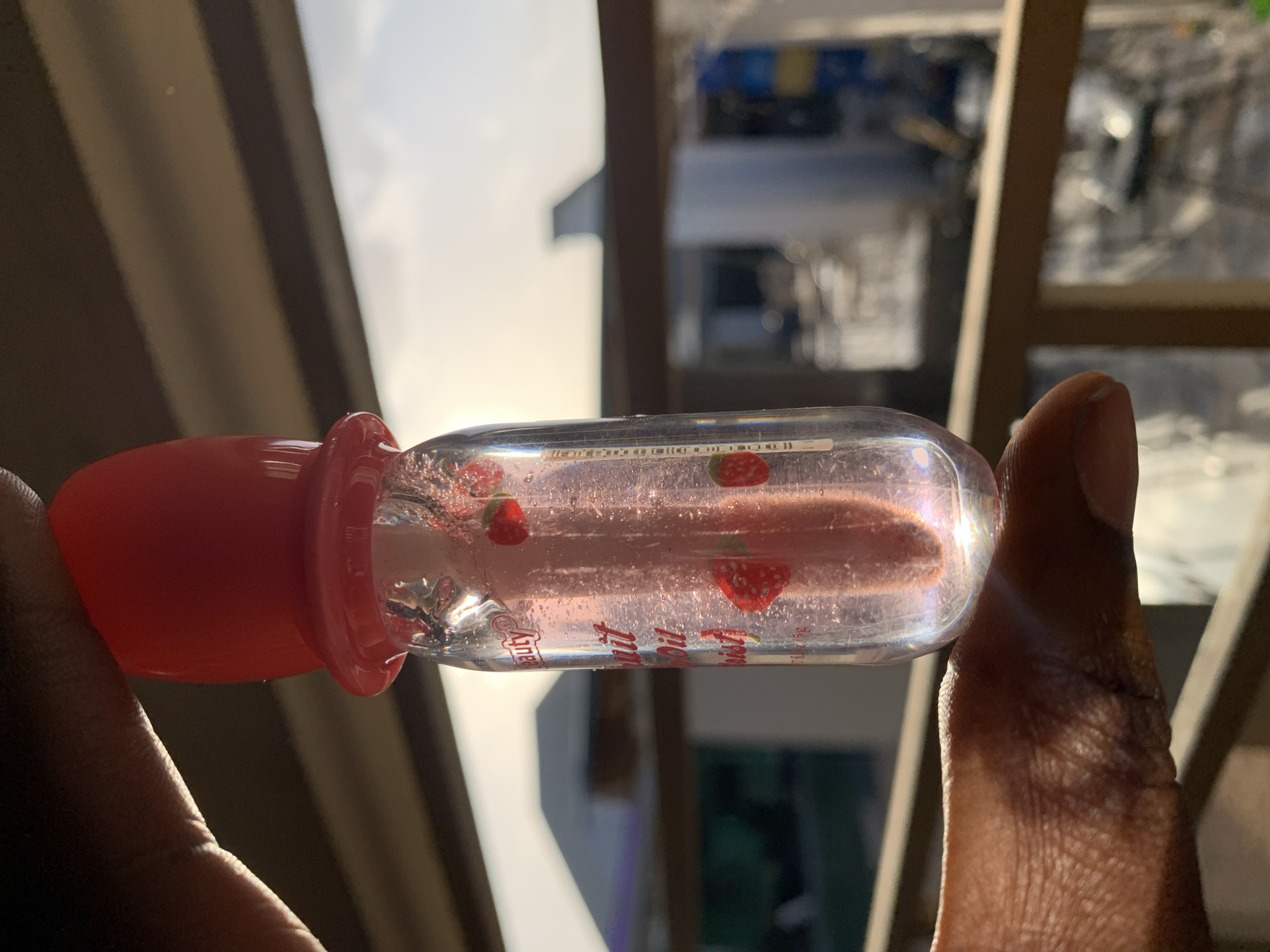 A person is holding a lip gloss tube against a blurred urban backdrop in the vicinity of New Stadium, Nigeria. The lip gloss has a clear body filled with liquid, scattered with strawberry decals, and a red cap. The person's fingers are visible holding the tube. Buildings and other urban structures are visible in the background, obscured by depth of field. Sunlight is present, creating highlights on the tube and the person's hand.