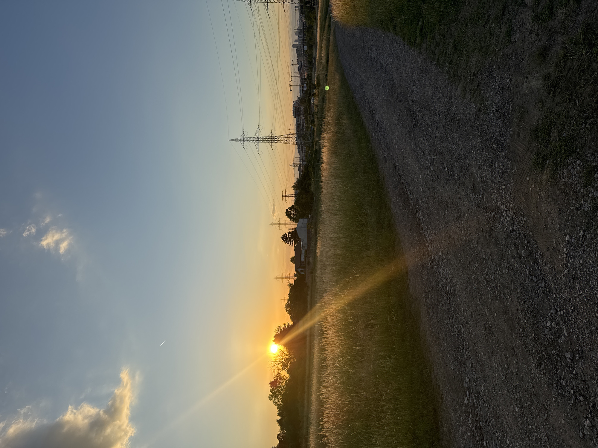 A gravel path curves through a grassy field towards a distant, hazy cityscape at sunset. The sun, a bright orb partially obscured by trees, casts golden rays across the scene. Tall power line towers and construction cranes dot the horizon. The sky is a gradient of soft blue and orange, with wispy clouds. There are no people visible in the image. The environment suggests an urban fringe or an undeveloped area on the outskirts of a city.