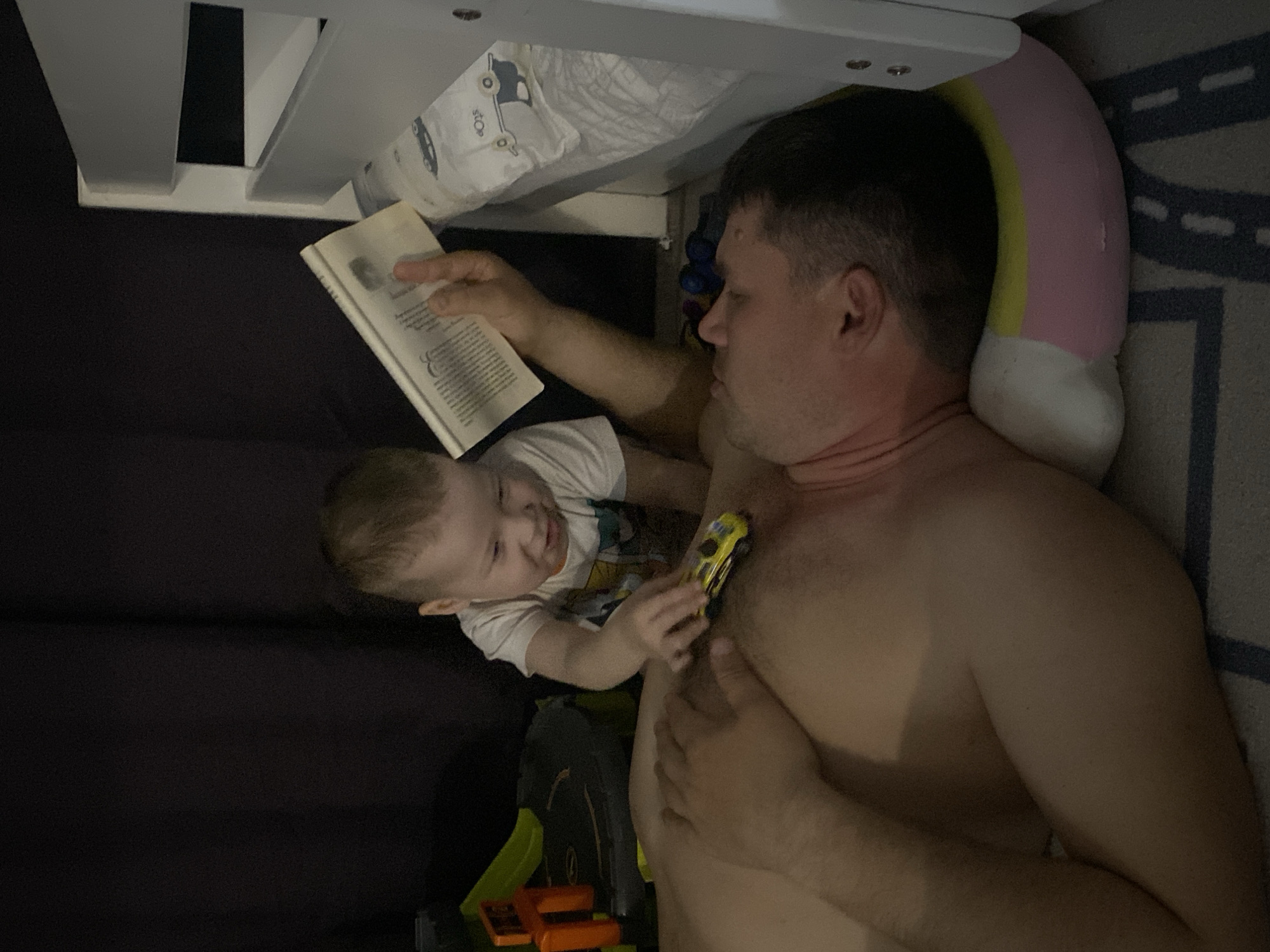 A man is lying down, propped up by a pillow, and reading a book. A young child is leaning on him and playing with a yellow toy car. A toy race track sits on the floor near them. The setting appears to be a bedroom. There is no information given in the image indicating its location. Therefore, it's impossible to tell if the image was taken in Sochi, Russia.