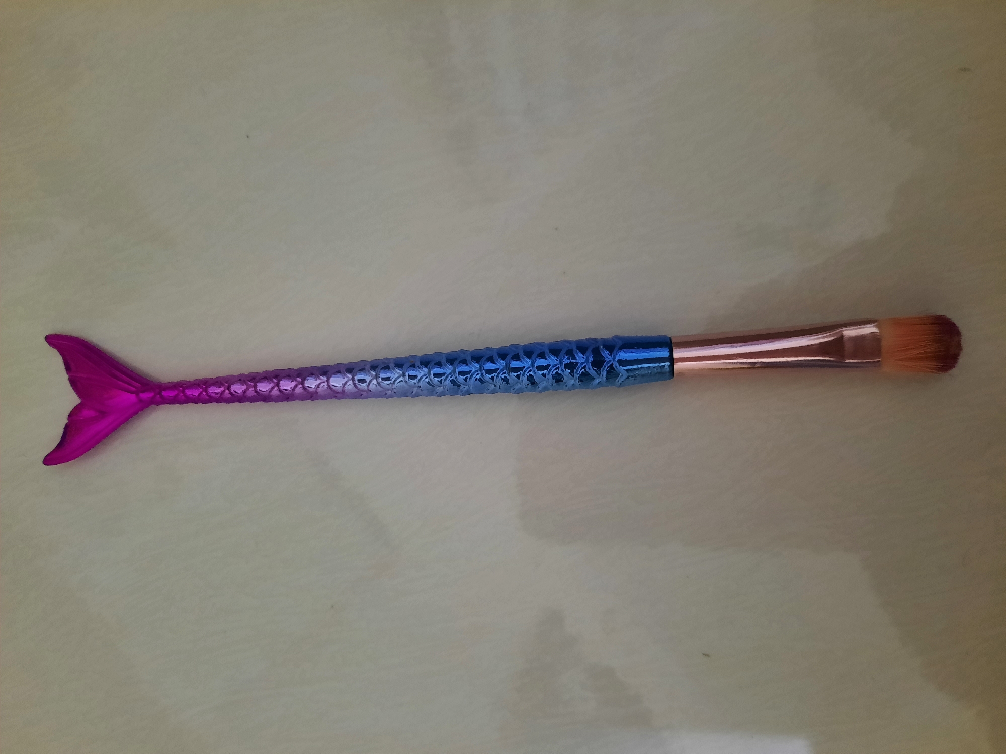 A makeup brush with a mermaid tail-shaped handle and pink bristles lies on a textured, light-colored surface. The handle is metallic, with a gradient of colors from pink at the tail to blue at the base of the bristles. The surface appears to be a countertop or table. The image does not contain information about the city of Lhokseumawe, Indonesia.