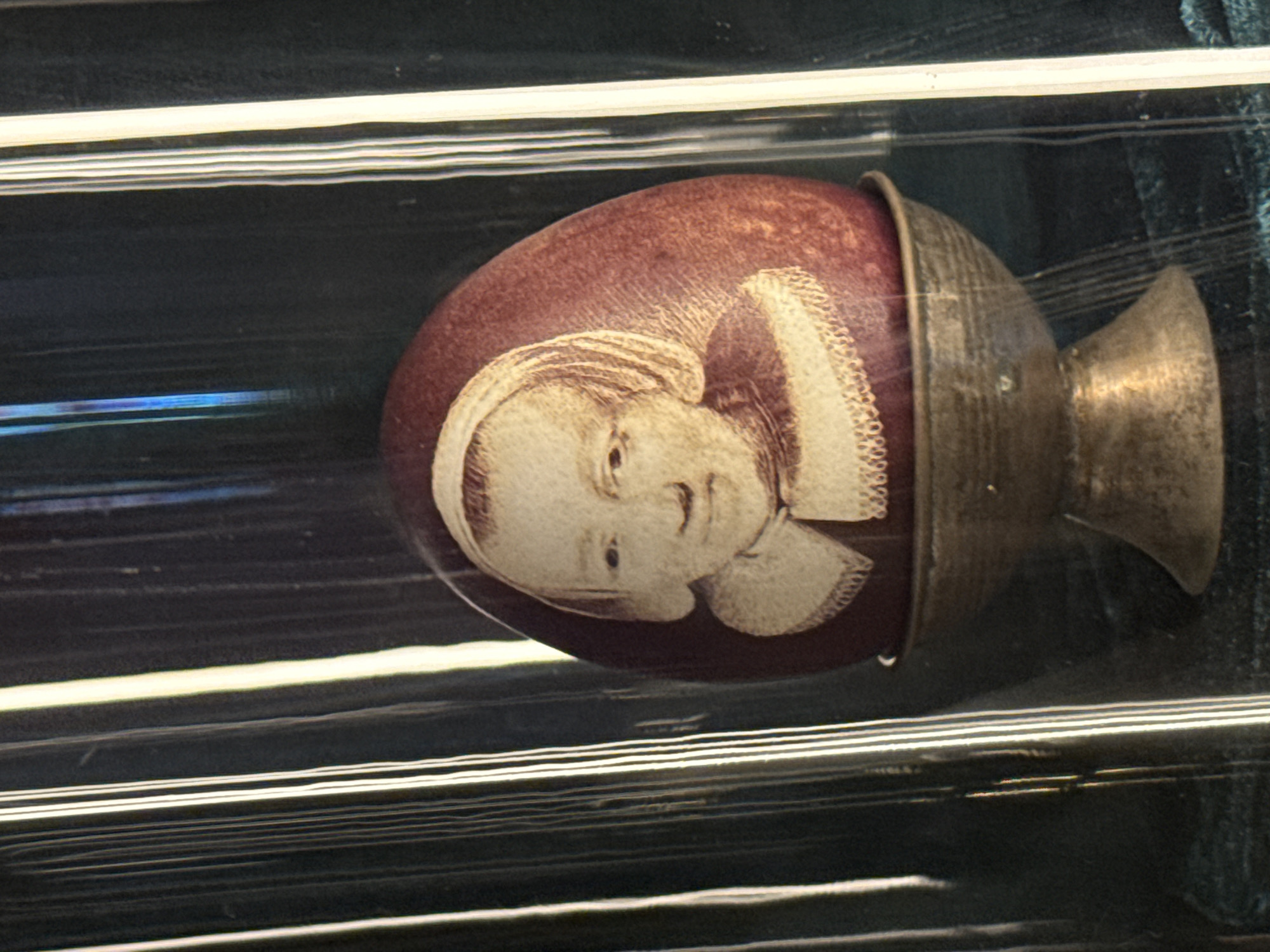 The image shows a red egg, decorated with a white portrait of a woman with a ruff collar, supported by a metallic base and stem. It is displayed within a protective case featuring horizontal bars, likely in a museum or exhibition setting in Chynadiiovo, Ukraine.