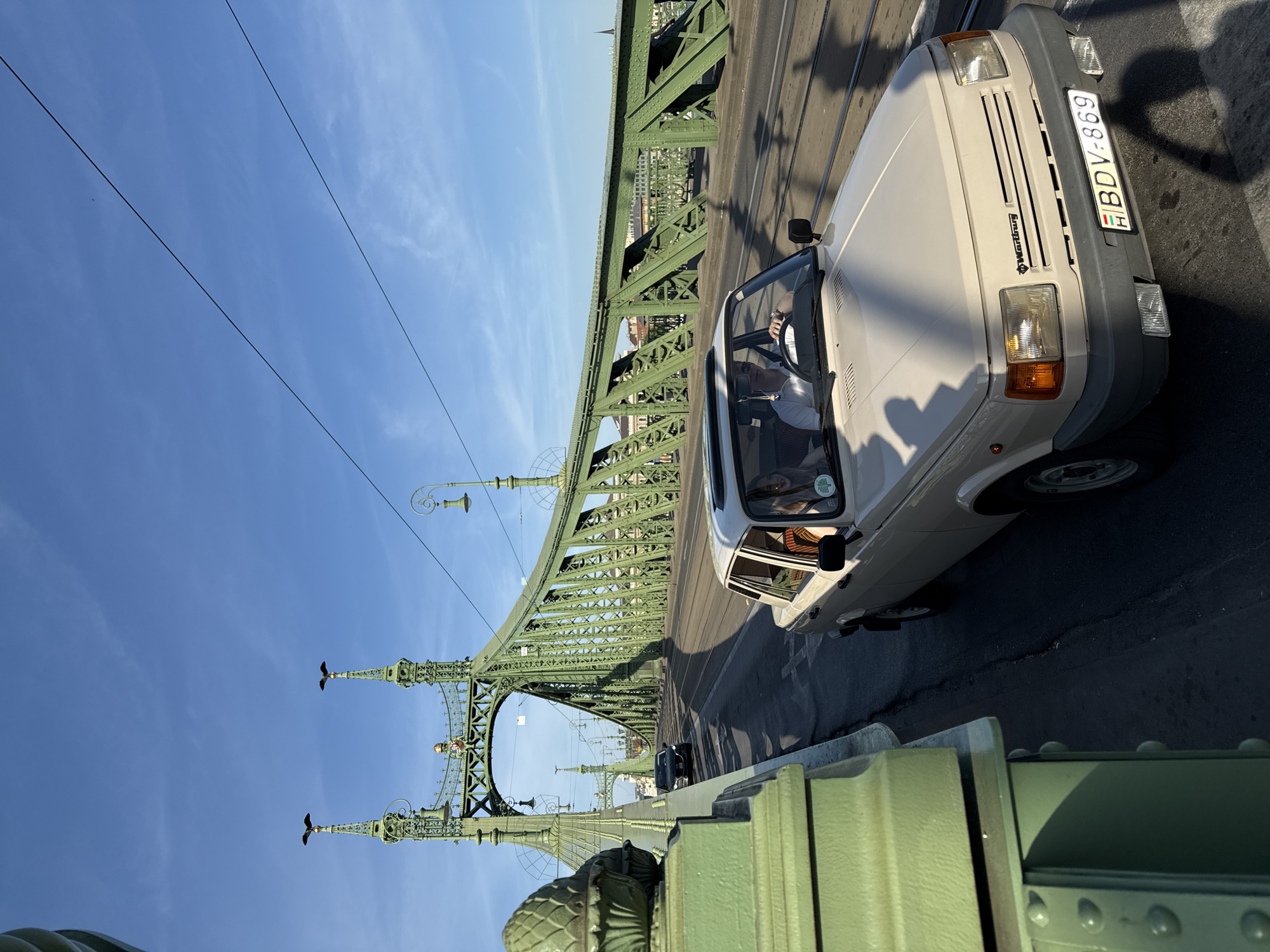 A light-colored Wartburg car is driving on the Liberty Bridge in Budapest, Hungary. The bridge's green steel structure is prominently displayed, with decorative turrets visible against a clear blue sky. There are people in the car. The car's license plate reads "BDV-869." The bridge roadway is asphalt.