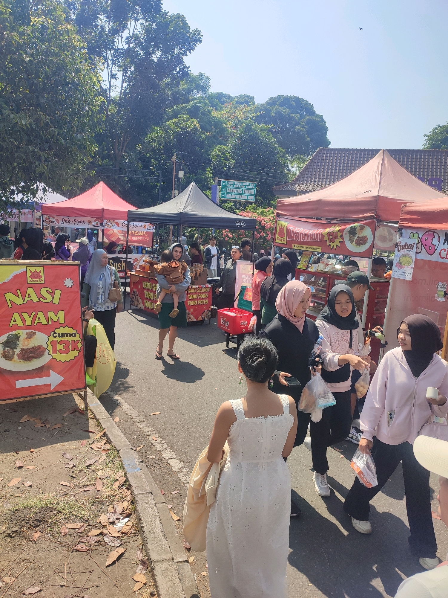 The image depicts an outdoor food market in Depok, Indonesia, likely near a university campus given the signage. A bright red banner advertising "Nasi Ayam" (chicken rice) for 13K is prominently displayed on the left. Several food stalls with colorful awnings are set up along a street. People, many wearing hijabs, are browsing the stalls and walking through the market. In the foreground, a woman in a white lace dress is walking away from the camera, while others are interacting at a stall selling drinks and snacks. The environment is sunny with trees and buildings visible in the background.
