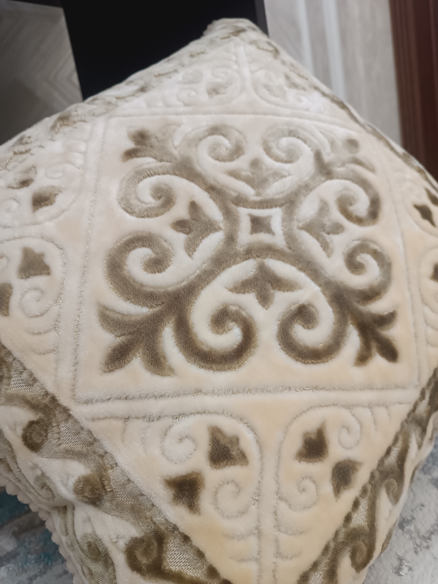 A close-up, high-angle shot shows a cream-colored, plush pillow with an embossed intricate pattern. The pattern features swirling, curvilinear motifs in a darker, brown hue, resembling floral or paisley designs. The texture of the pillow appears soft and velvety, with a subtle shimmer from what might be metallic threads woven into the fabric. The pillow is angled, with the top portion receding into the background, and it rests on a surface that is partially visible at the bottom of the frame, showing a blurred, greyish pattern. The background is dark and out of focus, suggesting an indoor setting, possibly a room in a house in Myrza-Ake, Kyrgyzstan. No people or actions are depicted.