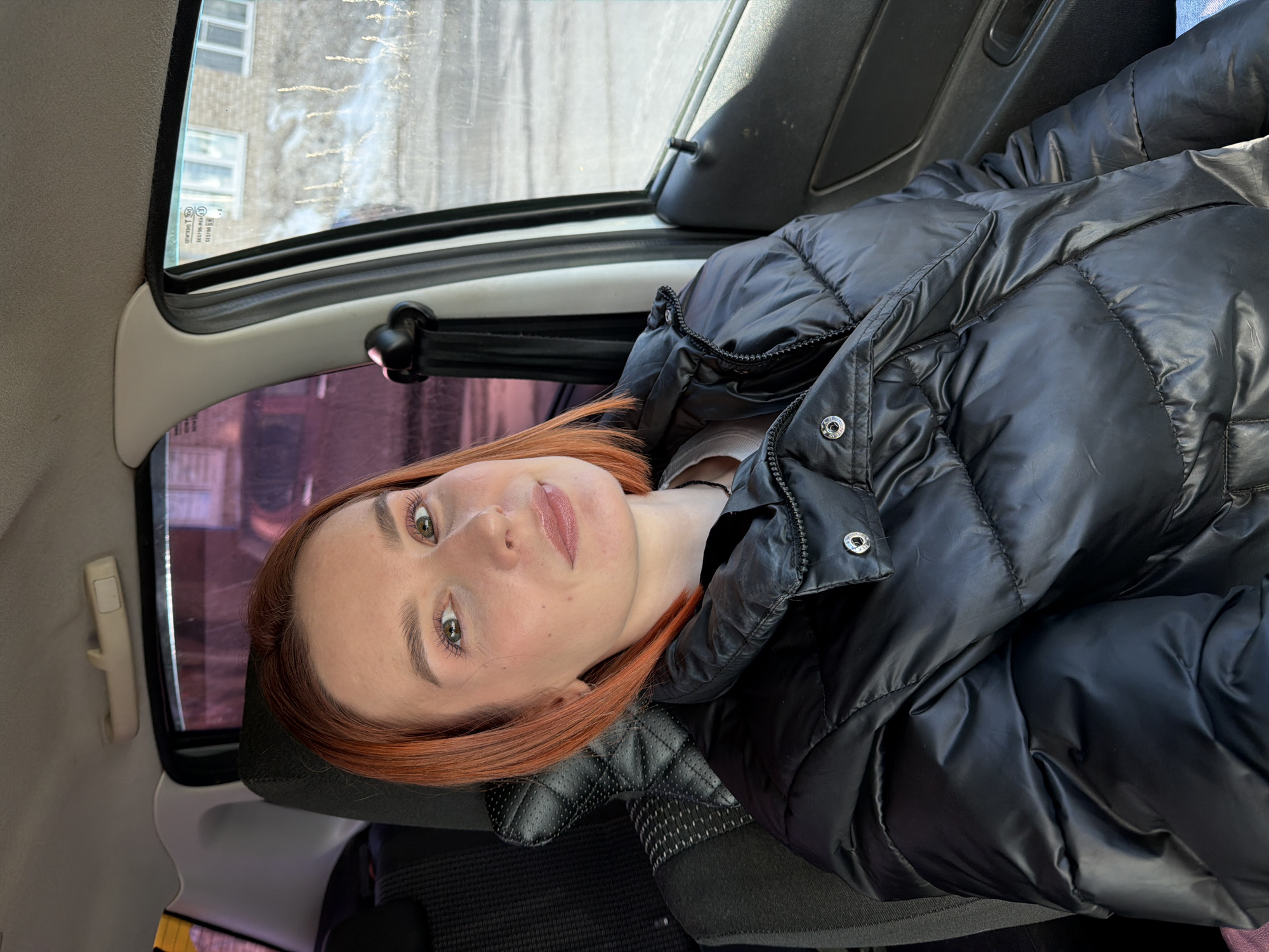A female subject, appearing to be in her 20s or 30s, is seated in the front passenger seat of a vehicle. She has shoulder-length reddish-orange hair, light skin, and green-brown eyes with visible light-colored eyeshadow. She is wearing a shiny black puffer jacket over a light-colored top and a dark beaded necklace, and her lips show a subtle pink tint. The subject is looking directly at the camera with a slight smile.
The vehicle interior features a dark, possibly quilted, seatback, a light-colored headliner with a grab handle visible above the window, and a dark grey door panel. The window on the left side of the frame displays a reddish-purple tint or reflection. Through the right side window, a multi-story residential building with white-framed windows is visible. Patches of snow or ice are present on the ground outside, and vertical dirt streaks are evident on the exterior of the window glass.
The architectural style of the visible building and the presence of snow are consistent with urban environments in Russia. However, a precise location within the city of Chaikovsky, Russia, is not discernible from the provided imagery.