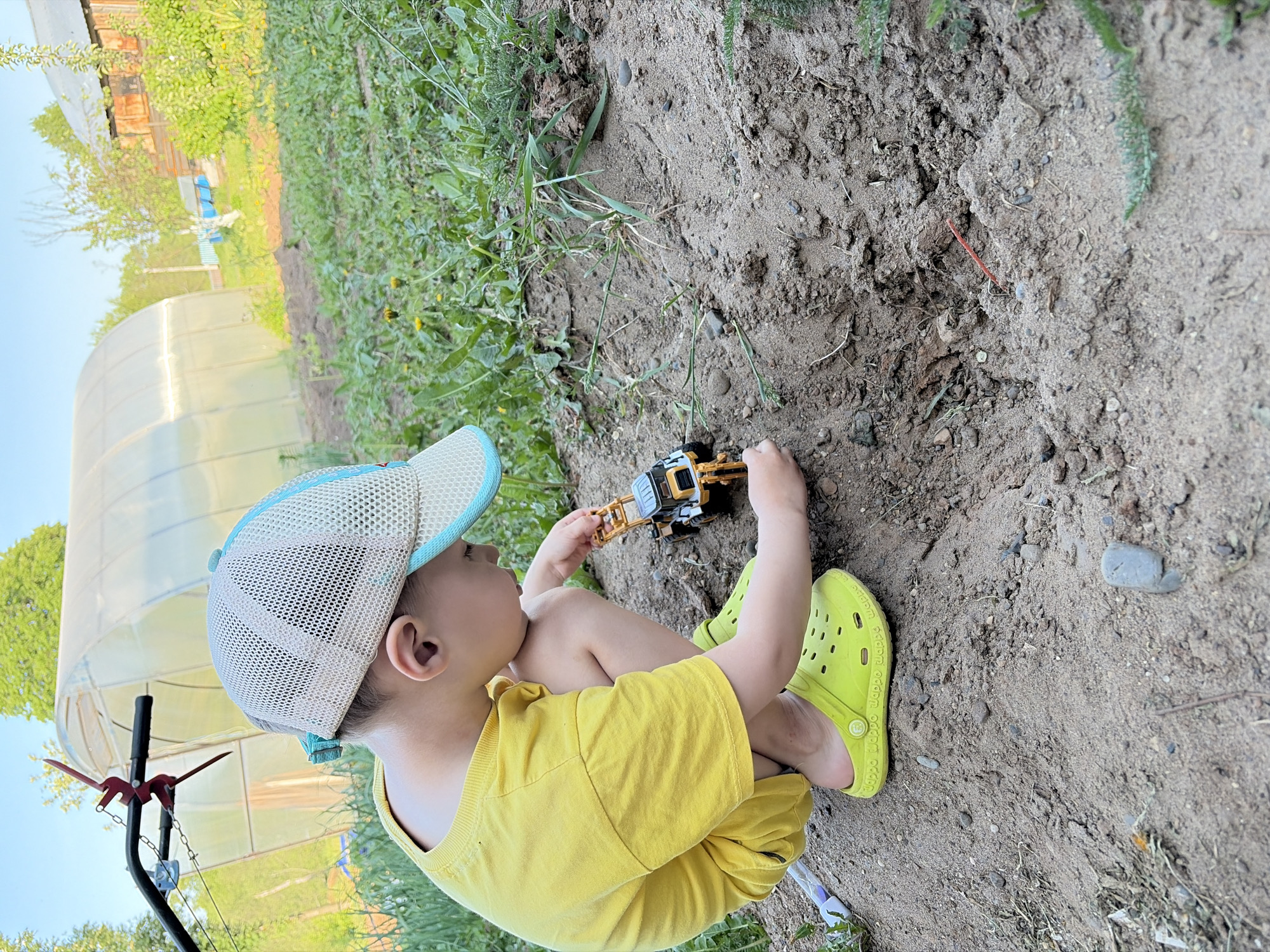 A child wearing a yellow shirt, yellow shorts, a white and blue baseball cap, and yellow Crocs is sitting on the ground, playing with a yellow and black toy bulldozer. The ground appears to be a mix of dirt and sand. Some vegetation is visible along the edges of the ground. A greenhouse structure is visible in the background. Based on the prompt, the location is presumed to be Чумна, Russia, although there are no visual cues to confirm this.