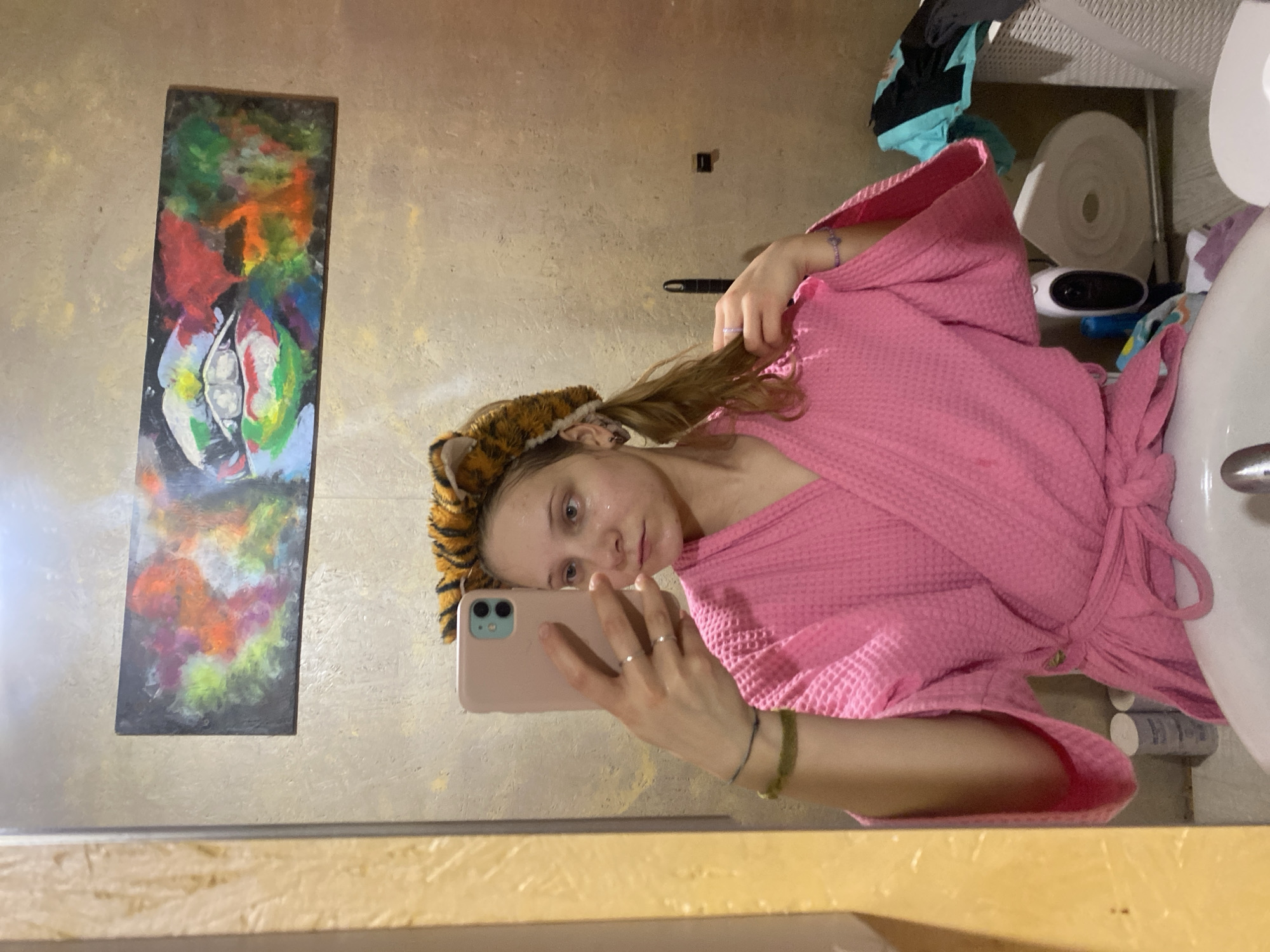 The image depicts a young woman in a pink bathrobe taking a selfie in a bathroom. She is wearing a tiger-striped headband. A painting hangs on the wall behind her. The bathroom contains a toilet and a sink. The location is in the city of Chaikovsky, Russia.
