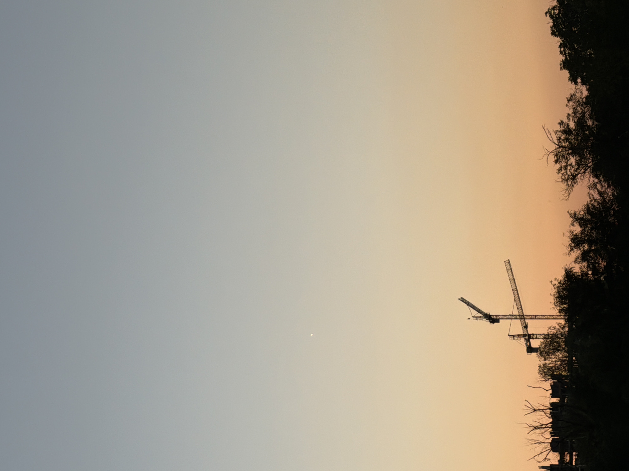 The image depicts a serene sunset sky over Plovdiv, Bulgaria, characterized by a gradient of soft blues and oranges. Silhouetted against this colorful backdrop are two towering construction cranes, their metallic structures stark against the fading light. Below the cranes, the dark outlines of trees and what appears to be unfinished buildings create a dense, natural and urban foreground. A single, bright star is visible in the upper portion of the sky, adding a touch of celestial wonder to the scene. The overall atmosphere is one of quiet anticipation, with the construction cranes suggesting ongoing development within the city.