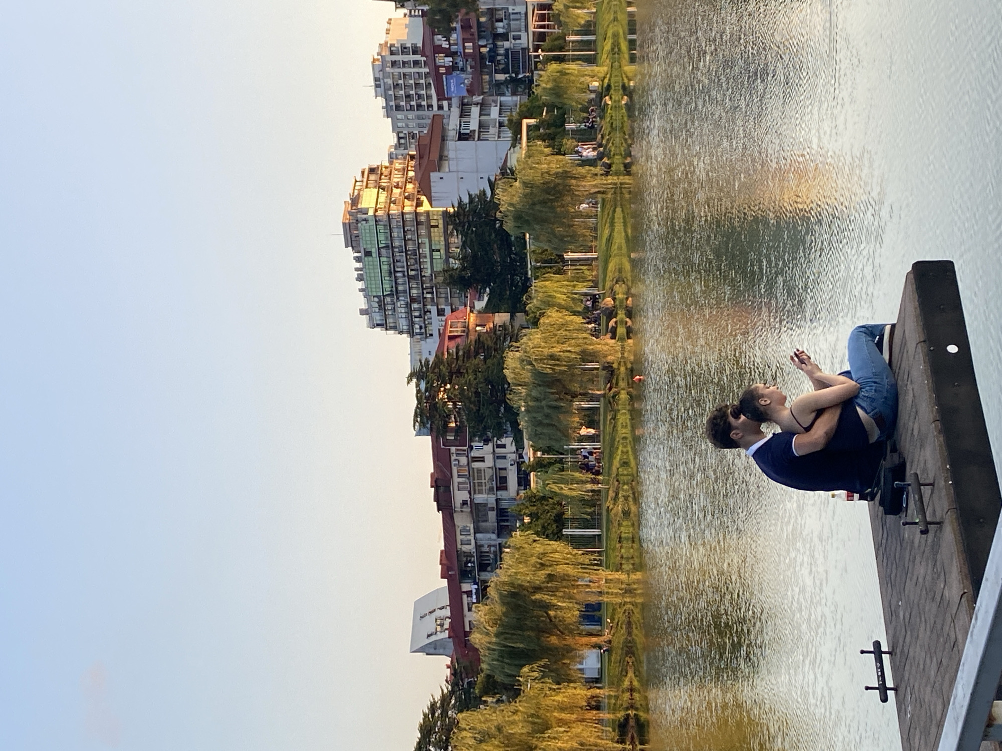 The image depicts a serene waterside scene in Batumi, Georgia, likely in the evening, given the warm lighting. In the foreground, a young couple sits closely together on a wooden pier extending into a calm body of water. The man, wearing a dark polo shirt and blue jeans, has his arm around the woman, who is dressed in a dark tank top and blue jeans. She is holding a smartphone and appears to be looking at it. A bottle of soda is placed on the pier near them.
The water reflects the soft hues of the sky and the surrounding environment, creating a shimmering effect. Beyond the pier and across the water, a lush green park area is visible, lined with tall, weeping willow trees whose foliage is illuminated by the setting sun. In the background, a line of multi-story buildings, characteristic of urban architecture, rises against the clear, pale blue sky. The buildings display a variety of architectural styles and colors, with some featuring balconies and numerous windows. The overall impression is one of peaceful coexistence between nature and urban development in the city of Batumi.