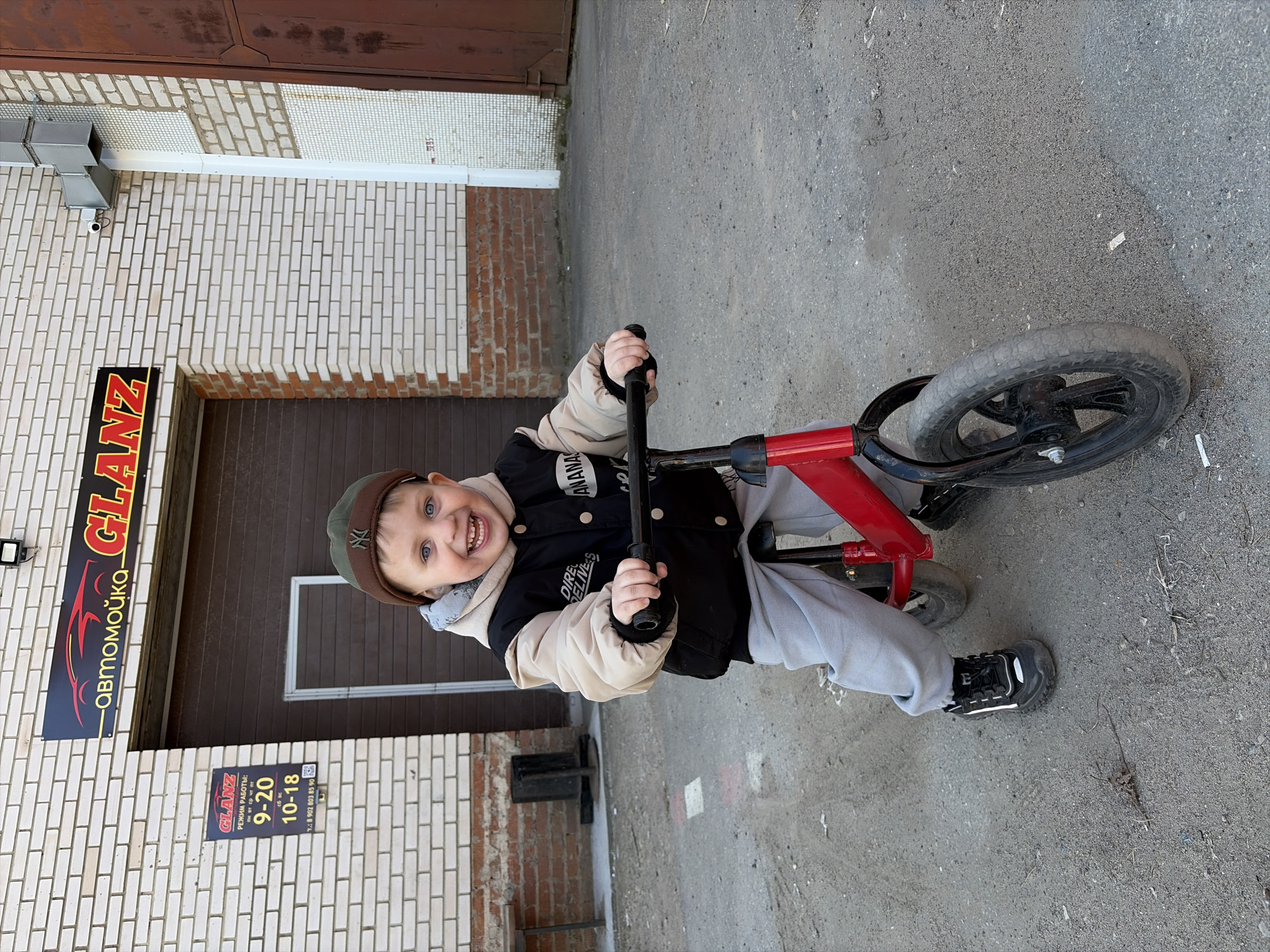 A young boy is riding a red balance bike in front of the Glanz car wash in Chaikovsky, Russia. The boy is wearing a cap, a jacket over a hooded sweatshirt, and grey sweatpants with black and white sneakers. He is smiling and looking at the camera. The Glanz car wash sign is visible on the brick building behind him, along with the business hours posted below it. The ground is grey concrete.