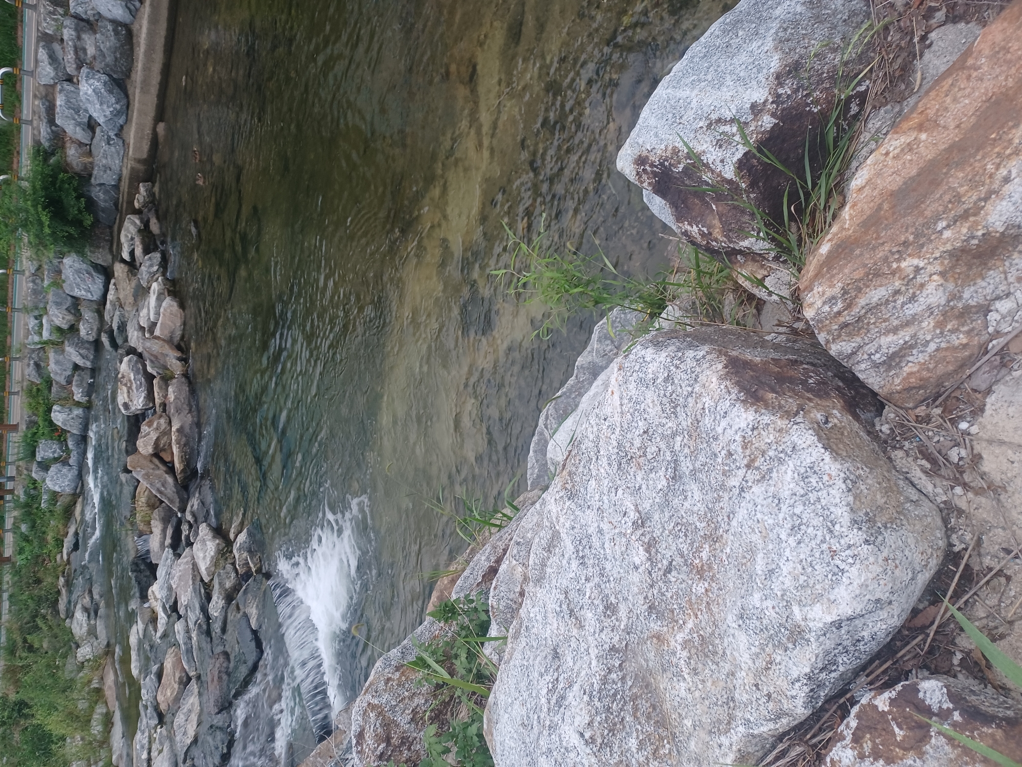 The image depicts a natural stream with rocky banks in Seoul, South Korea. The water flows over smooth stones and a small waterfall is visible on the left. The banks are lined with large boulders and some greenery. In the middle ground, two ducks can be seen swimming in the water. The background shows more rocks and vegetation, with a paved path and railings visible in the upper right corner. The overall scene is tranquil and scenic.