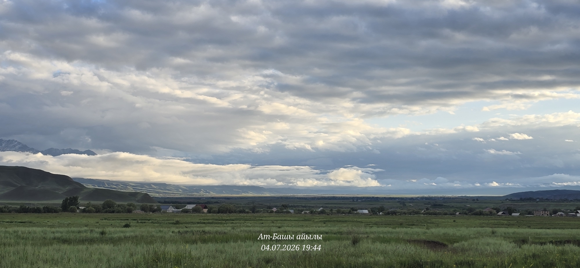 The image depicts a wide, expansive landscape under a dramatic, cloudy sky. In the foreground, a vast field of lush green grass stretches towards a line of trees and scattered houses, indicative of a rural settlement. The village appears to be situated in a valley, with rolling hills and mountains forming a backdrop. The mountains in the distance are partially obscured by clouds, and the peaks of some appear snow-capped. The lighting suggests it is late afternoon or early evening, with a warm glow illuminating some of the clouds. Text overlays in a non-English script and a date "04.07.2026 19:44" are present in the lower right quadrant of the image. The location is identified as At-Bashy, Kyrgyzstan.