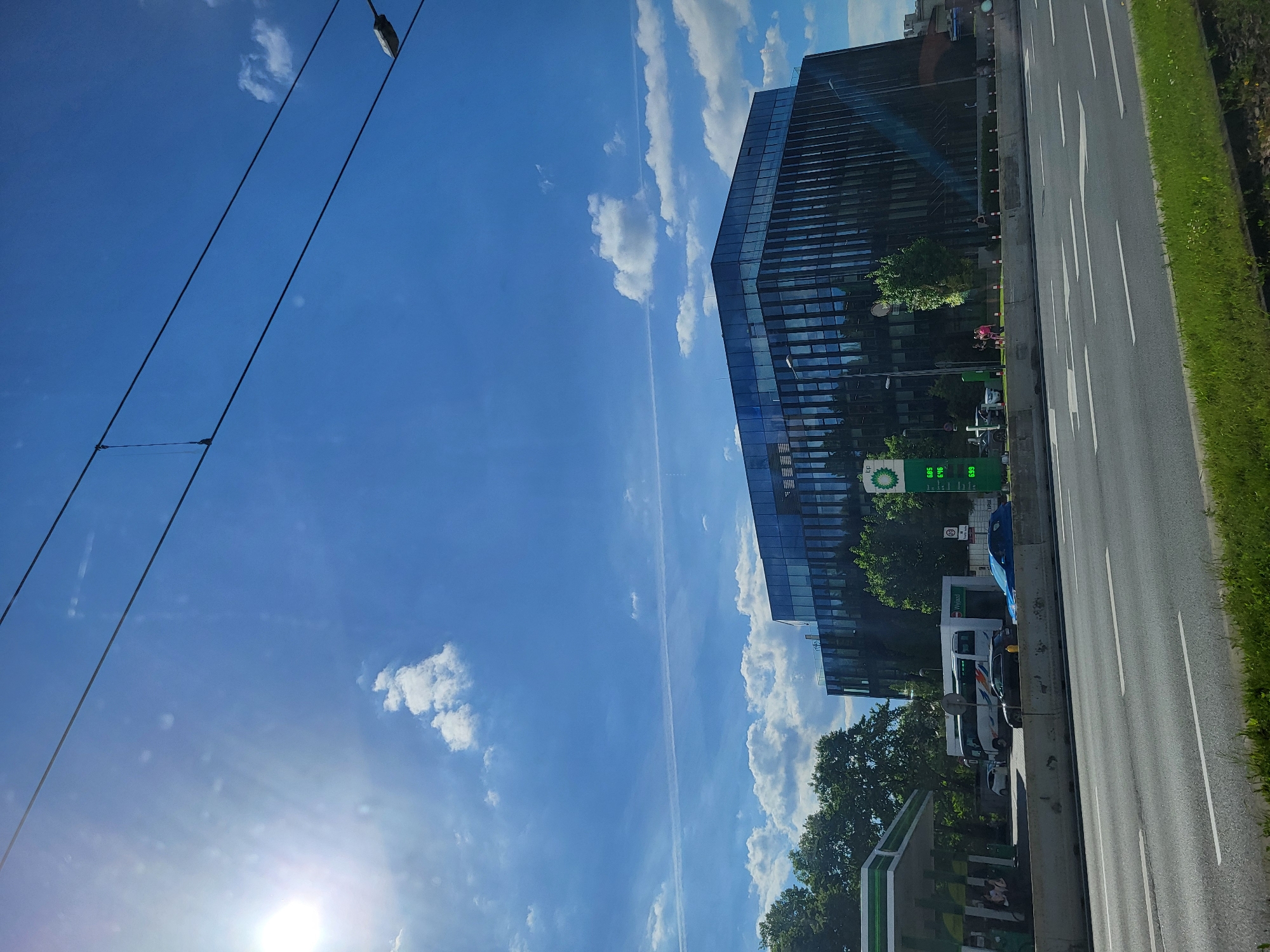 The image depicts a view in the city of Unknown, Unknown, showing a modern glass office building, a BP gas station, a roadway, and a bright blue sky with scattered clouds.
The building is tall and constructed with reflective glass, reflecting the sky. A BP gas station is adjacent to the building, with visible price signage. A multi-lane roadway runs alongside the gas station.
Overhead wires are visible in the sky. The sun is also in the image.