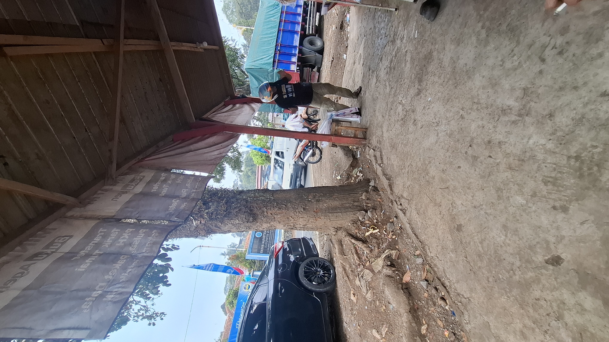 The image captures a street scene in Cilaku, Indonesia, likely near a roadside stall or shelter. In the foreground, a large tree with exposed roots stands prominently. To the left, a black sedan is parked on a dirt and gravel area. In the midground, a man wearing a black t-shirt, camouflage pants, and a helmet is standing and holding something. A motorcycle is parked behind him, and a wooden stool with plastic bags on it is next to him. Further back, a white van is driving on a road, and a large blue truck with a green tarp-covered load is parked. Two blue flags with text are visible in the background, one of which has the words "BADAN NARKO", suggesting a government agency presence. The ground is a mixture of concrete and dirt. The overall environment appears to be a public area with vehicle traffic and pedestrian activity.