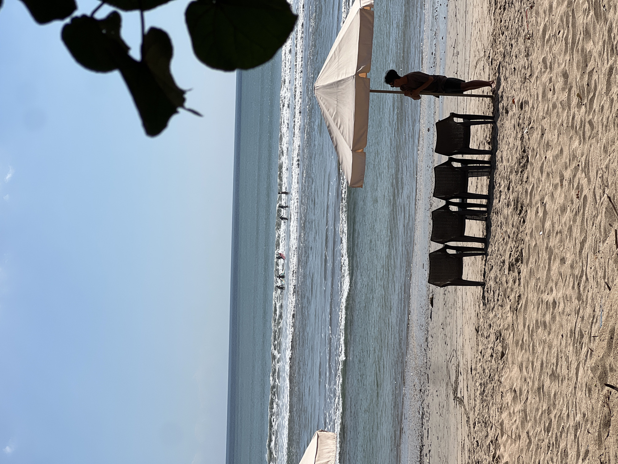 A view of the Kuta beach in Bali, Indonesia, under a bright blue sky. The foreground is dominated by sand, with some debris scattered across it. Four dark plastic chairs are lined up on the sand, facing the ocean. To the right of the chairs, a person stands next to a light-colored beach umbrella, holding its pole. In the middle ground, the ocean displays gentle waves with white foam. Several figures, appearing to be surfers with surfboards, are visible in the water further out. The background consists of the vast expanse of the ocean meeting the horizon under the clear sky.