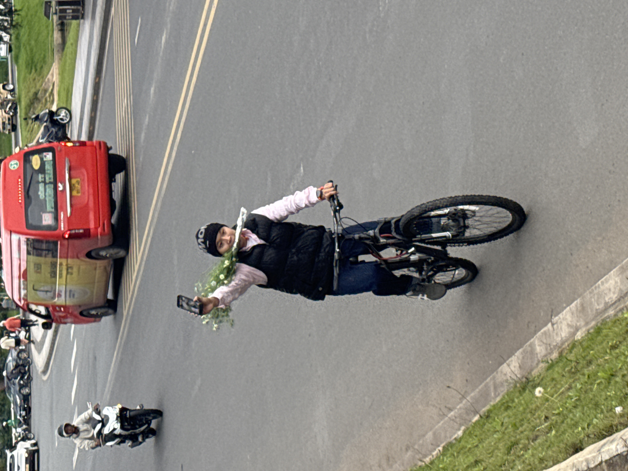 The image shows a person on a bicycle in Da Lat, Vietnam, holding a bouquet of flowers and a mobile phone. The person is wearing a black vest over a pink shirt, dark blue jeans, and a black beanie. They are riding on a paved road, with a grassy embankment to the left. In the background, there is a red van, several cars, and other motorbikes. The setting appears to be a street in a city.