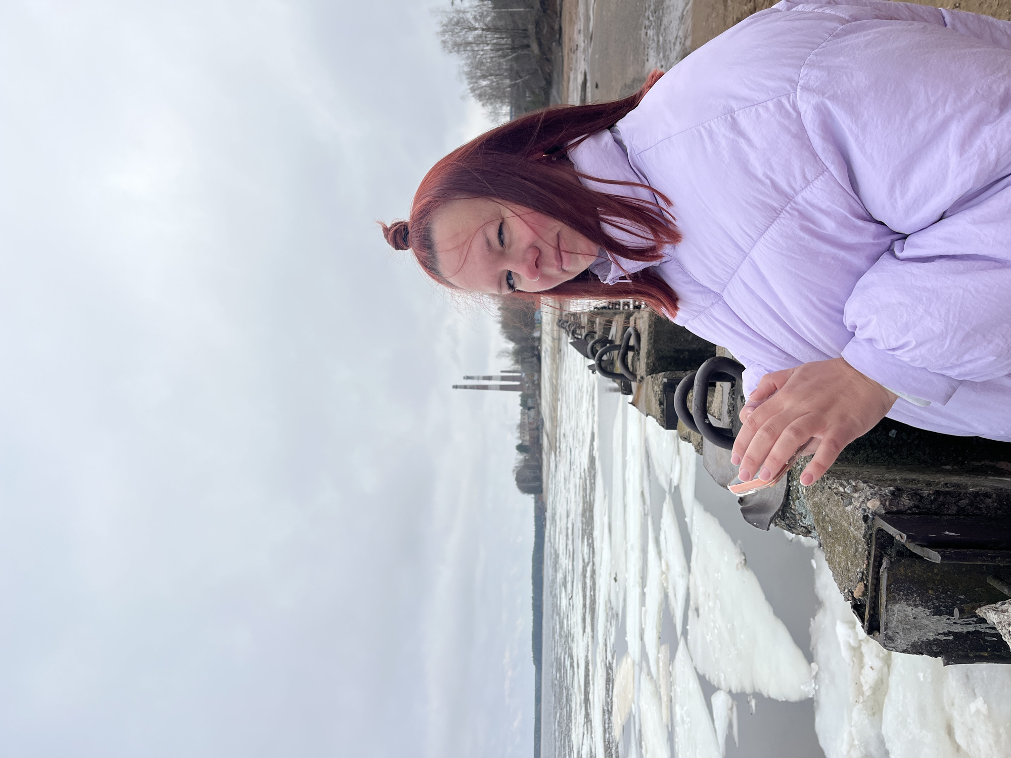 A young woman with reddish-brown hair is standing next to a concrete pier in Chaikovsky, Russia. She is wearing a light purple puffer jacket and has her hair tied up in a bun. The river is covered in ice floes, and in the distance, industrial chimneys can be seen. The sky is overcast.