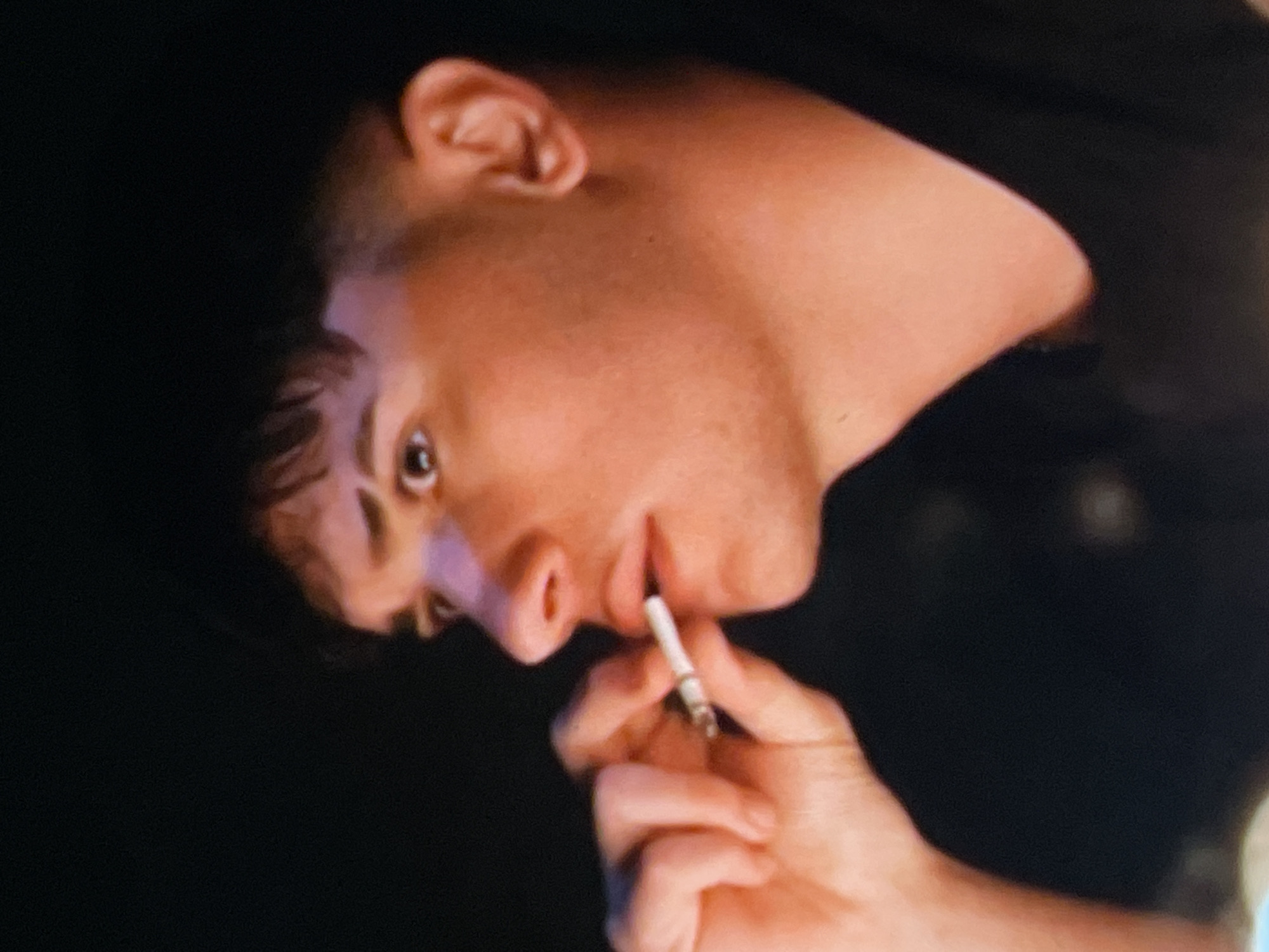 The image shows a close-up, profile view of a young man's face. He has dark, short hair with a faded cut and his eyebrows are dark and shaped. He is looking to the right with a slightly surprised or concerned expression. His mouth is slightly open, and he is holding a cigarette between his fingers, bringing it to his lips. The lighting is dim, casting shadows and highlighting his features with a warm, reddish tone. The background is dark and out of focus, providing no environmental context. There is no indication of Harmanli, Bulgaria.