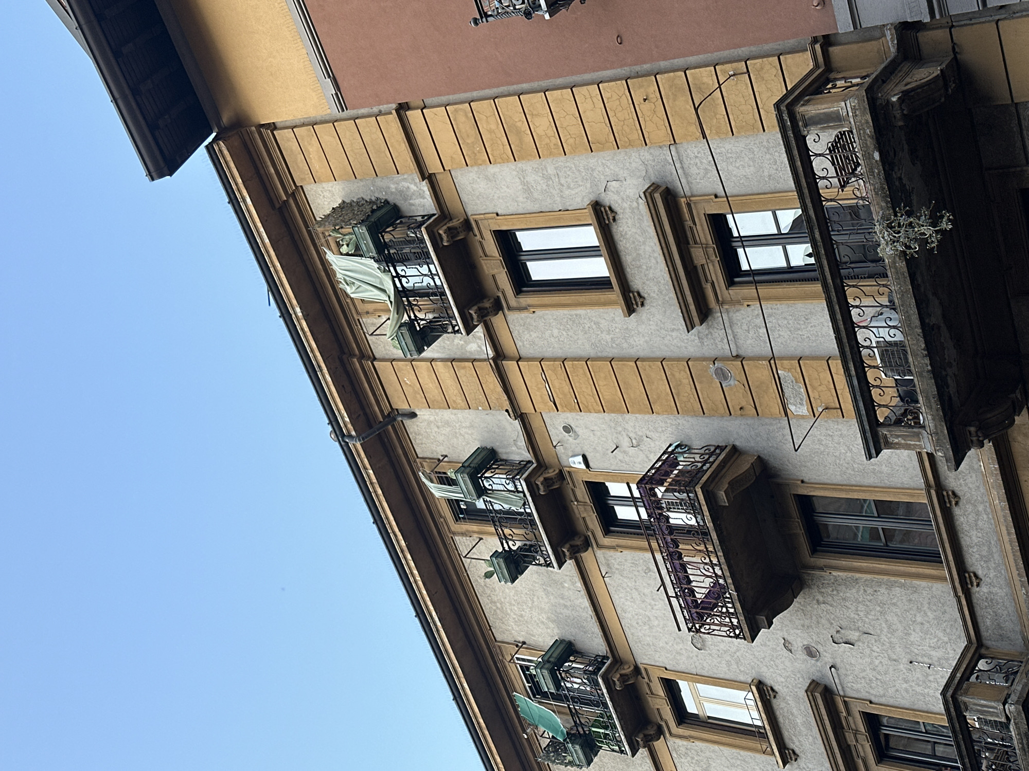 The image captures the façade of a building in Milan, Italy, presenting an upward-angled perspective. The building's exterior is characterized by a yellow trim contrasting with a gray plastered wall. Windows are arranged vertically, most featuring black frames and some accompanied by wrought iron balconies. Some balconies are adorned with plants or fabric coverings. The sky, a clear blue, is visible to the left of the building.