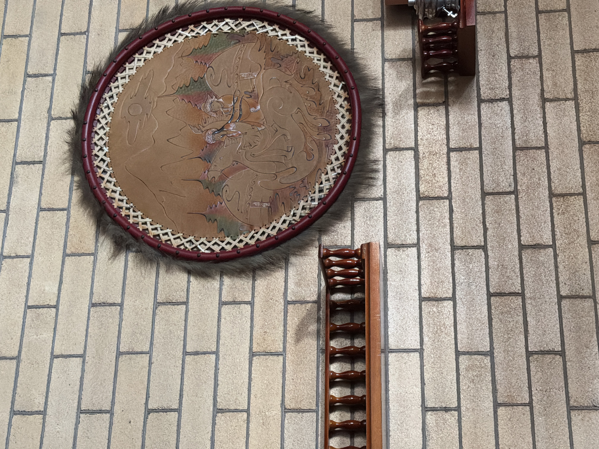 The image shows a circular, decorative item hanging on a brick wall. The item has a dark red rim and is covered with dark fur around the edge. Inside the rim, there is a design embossed on leather, depicting a scene with mountains, trees, and figures that appear to be on horseback. Below the circular decoration, there is a wooden shelf with a decorative railing. The brick wall is light-colored with dark grout lines. The image is taken from a medium distance, capturing the wall and the objects in detail. The setting appears to be indoors, possibly a home or a cultural establishment. There are no people visible in the image. The location of the city administration building in Qarağandı, Kazakhstan is not discernible from the visual information provided.