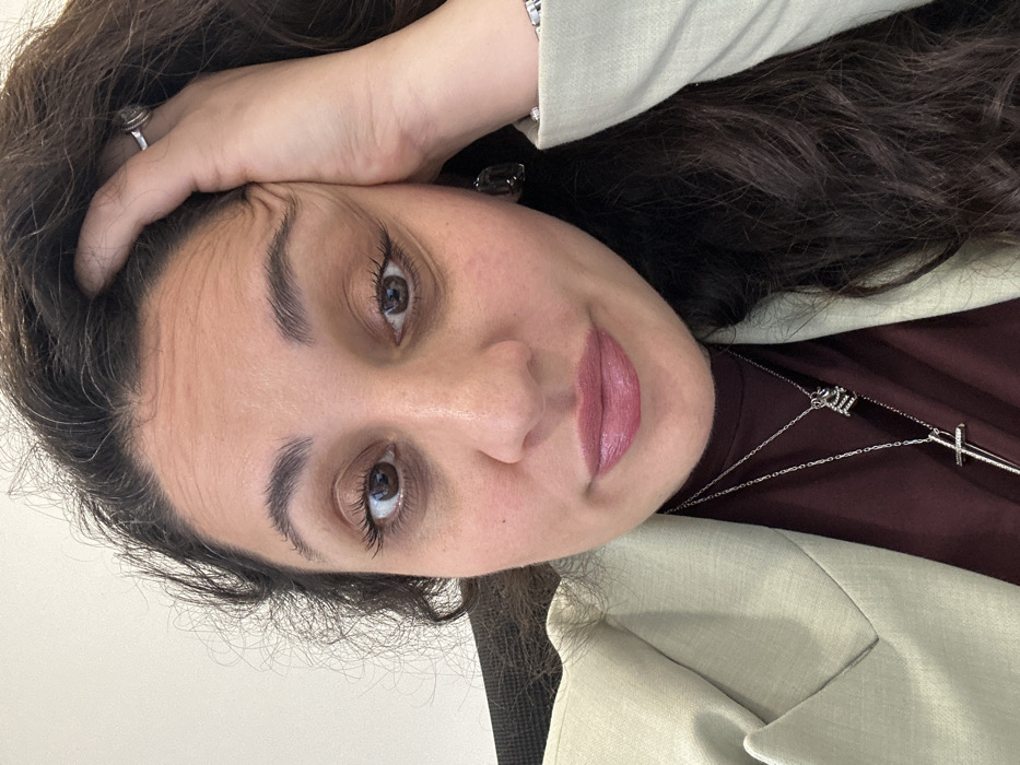 A woman poses, head resting on her hand, displaying a silver ring. She has brown eyes, full eyebrows, and wears pink lipstick. A silver necklace with a cross pendant adorns her maroon top. She is wearing a light-colored blazer. Her dark, wavy hair frames her face. Her skin tone is light to medium. The image is shot at an angle, lending a casual and approachable feel.