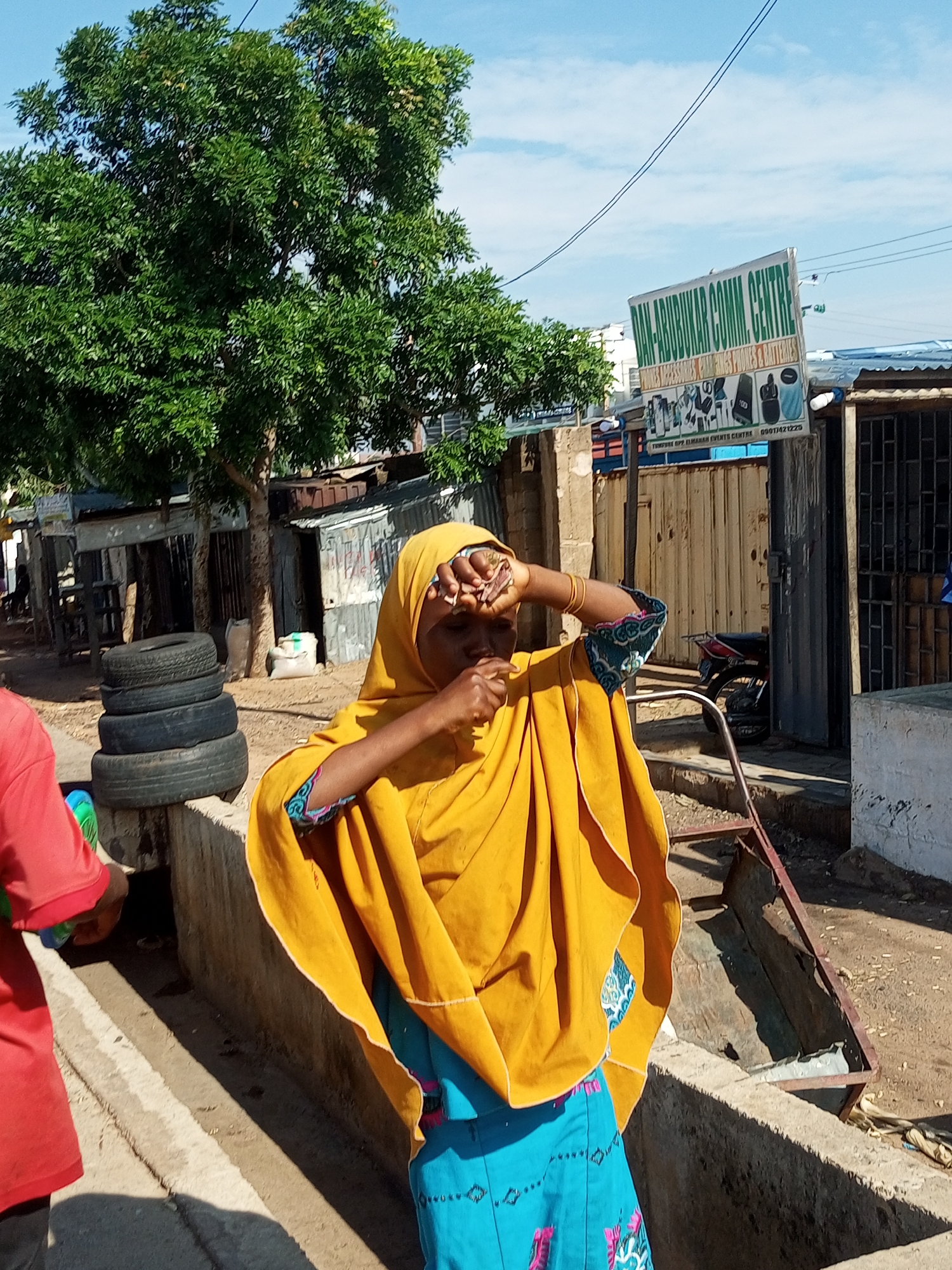 In Tempure, Nigeria, a young woman wearing a yellow hijab and a blue skirt stands in front of a building. She is shielding her eyes from the sun. To her left, a stack of tires is visible, and further back, corrugated metal structures line the street. A sign for "DAN-ABUBAKAR COMM. CENTRE" is mounted on the building. The scene suggests a bustling marketplace or commercial area in Nigeria.