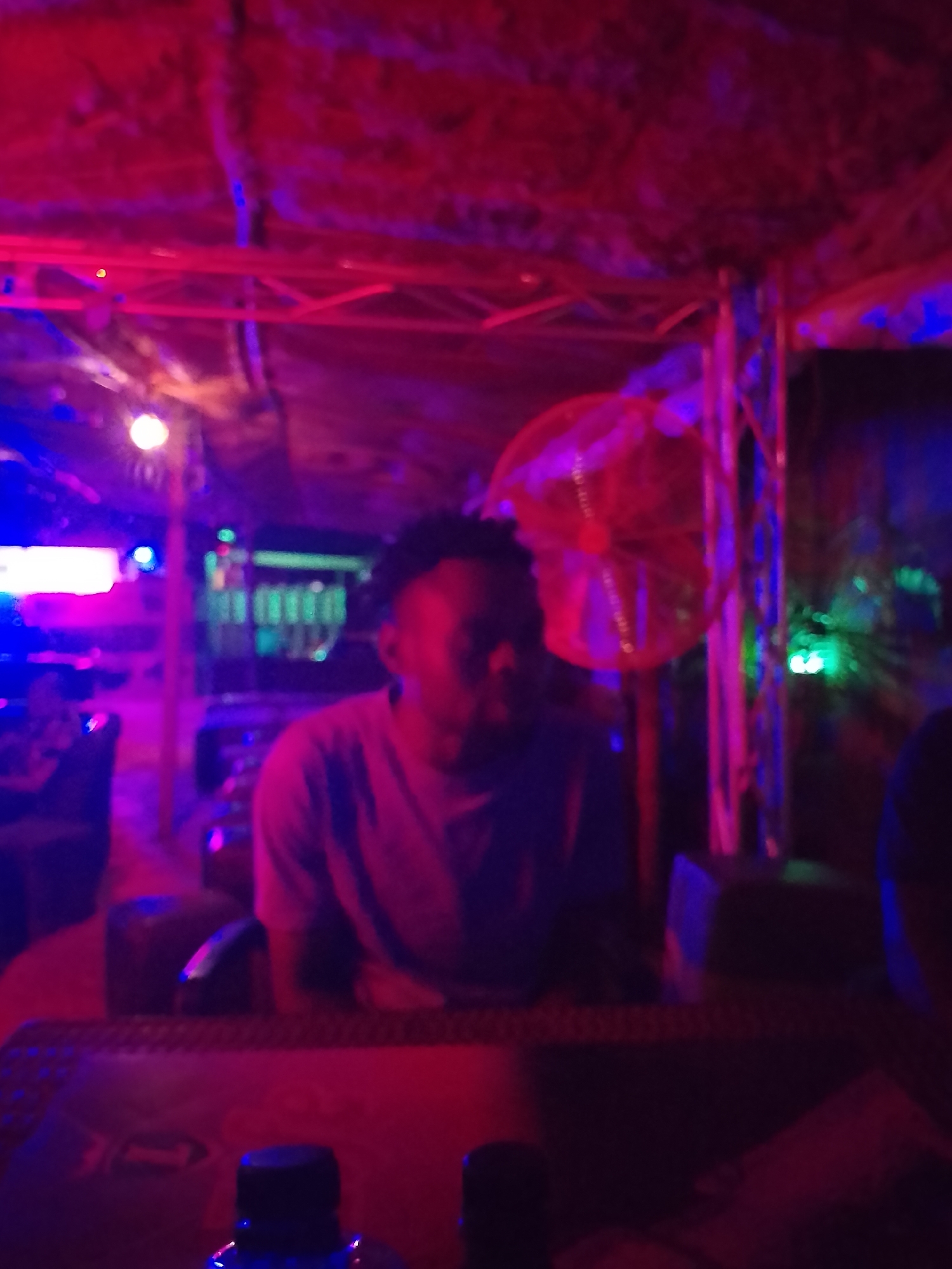The image depicts a dimly lit interior space in an unidentified location within the city of Unknown, Unknown. A person with dark hair is seated at a table, illuminated by red and blue lighting. A fan is visible in the background. Bottles are on the table in the foreground. There are other indistinct figures and objects scattered throughout the scene.