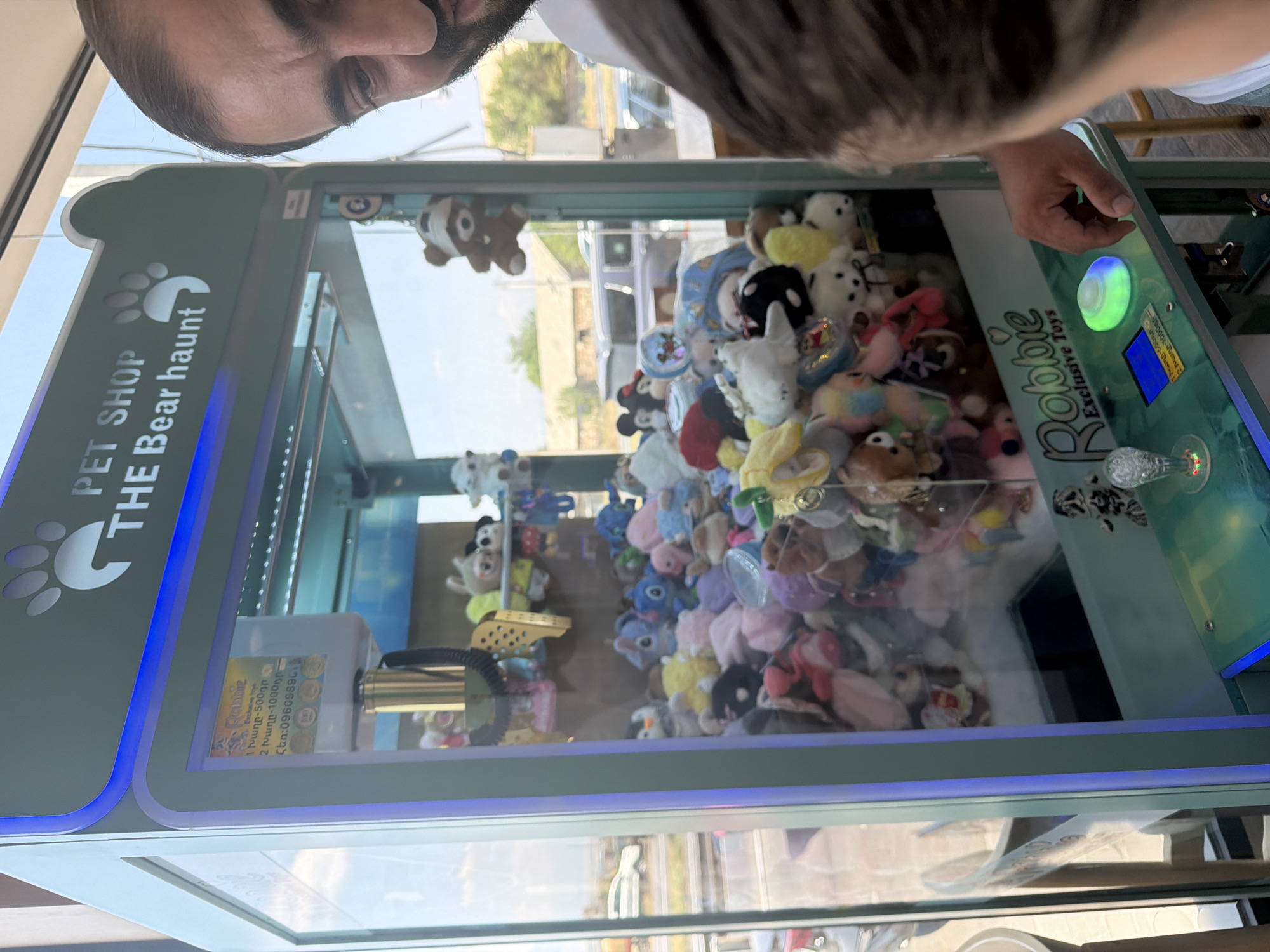 In Verin Ptghni, Armenia, a man is shown with his face partially visible on the right side of the frame, looking towards a claw machine filled with plush toys. The machine, labeled "PET SHOP THE Bear haunt," has a blue neon trim and a graphic of a paw print. On the top left of the machine, a colorful sign displays information about pricing, including "1 pound - 500t," "2 pound - 1000t," and a phone number. The claw machine's interior is packed with various stuffed animals, including a brown teddy bear hanging from the top. In the foreground, a child's head and shoulder are visible, also looking at the machine. The child's hand is reaching towards the illuminated control panel of the claw machine. The environment appears to be indoors with natural light coming from a window on the left, which shows a blurred view of cars and buildings outside.