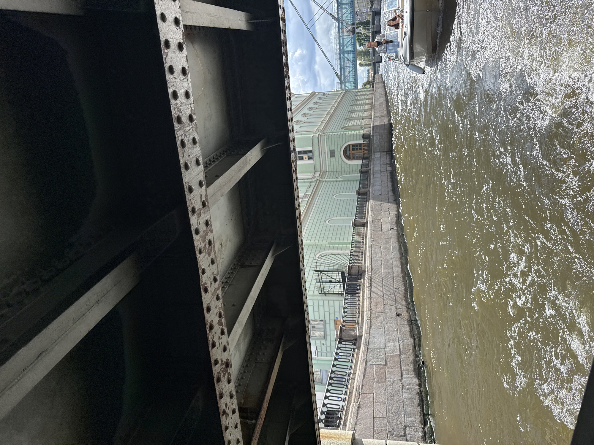 This image depicts a view from beneath a bridge, looking out over a canal in Saint Petersburg, Russia. The canal water is a murky brown, with sunlight reflecting off its surface, creating sparkling patterns. On the right bank of the canal, a light green building with classical architectural elements and arched windows is visible. A black metal fence with ornate railings lines the embankment in front of the building. Further down the canal, another bridge, this one appearing to be a suspension bridge with a blue structure, can be seen in the distance. A small motorboat with two people aboard is moving away from the viewer in the canal. The boat has a white hull and a bimini top. One person, a man wearing a light-colored jacket and dark pants, is standing at the helm. Another person, a woman wearing a dark top, is seated. The underside of the bridge, constructed of metal beams and rivets, frames the upper portion of the image. The overall scene suggests a daytime boat tour or passage through one of Saint Petersburg's many canals.