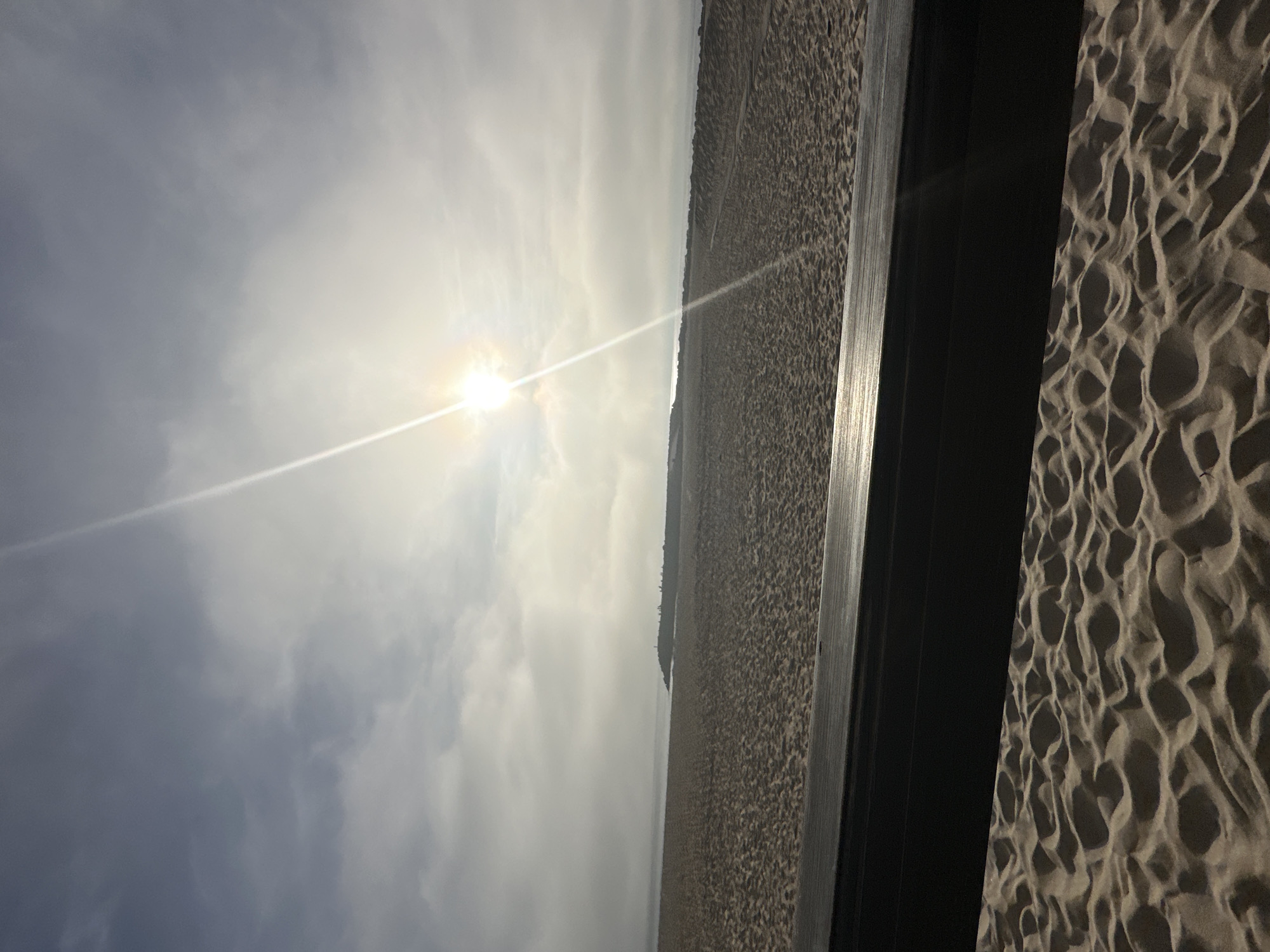 View of the beach and sky from a dark, possibly shaded area, potentially from a building or structure. The beach appears vast and sandy, stretching to the horizon. The sky is overcast with the sun shining through the clouds, creating a bright spot with a visible sunbeam. The city of Espinho is likely the location. There are no visible people.