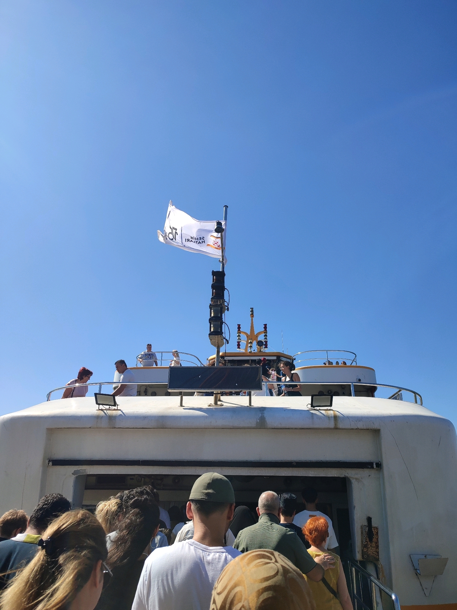 A group of people are boarding a ferry on a clear, sunny day. The ferry has a white exterior and a white flag with a logo on its mast. Several people are visible on the upper deck of the ferry, and more are queuing to board. The entrance to the ferry is an open doorway, and a set of stairs leads up to it. The scene appears to be taking place in the city of Kadıköy, Turkey, a major transportation hub on the Asian side of Istanbul, known for its ferry terminals connecting to the European side and other islands.