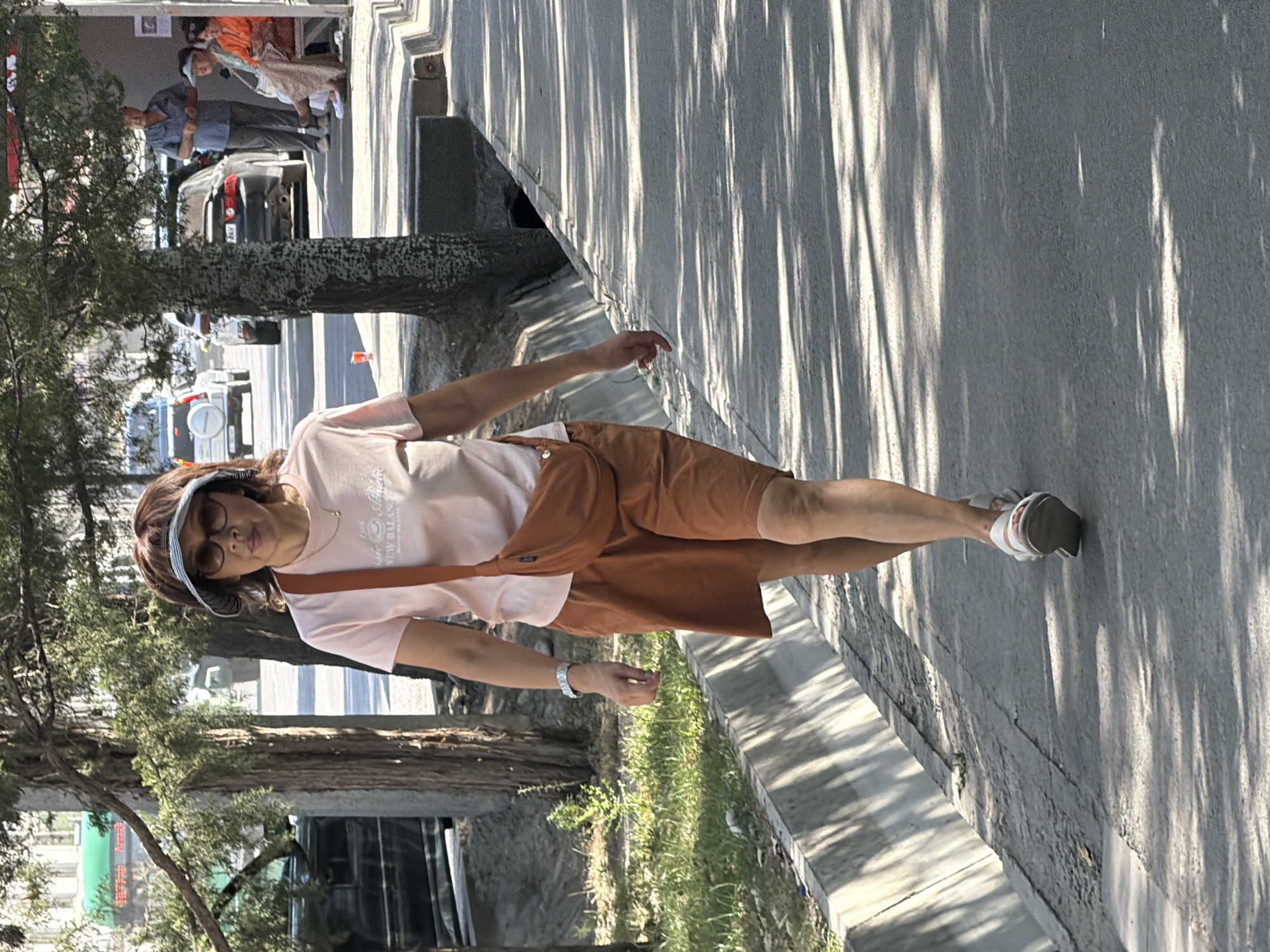 A woman with brown hair, wearing a pink t-shirt, brown shorts, white sandals, sunglasses, and a visor, walks along a sidewalk. She has an orange crossbody bag slung over her right shoulder. She is looking towards the camera. In the background, there are trees, cars, and other people sitting on a bench. The scene appears to be a street in a city.