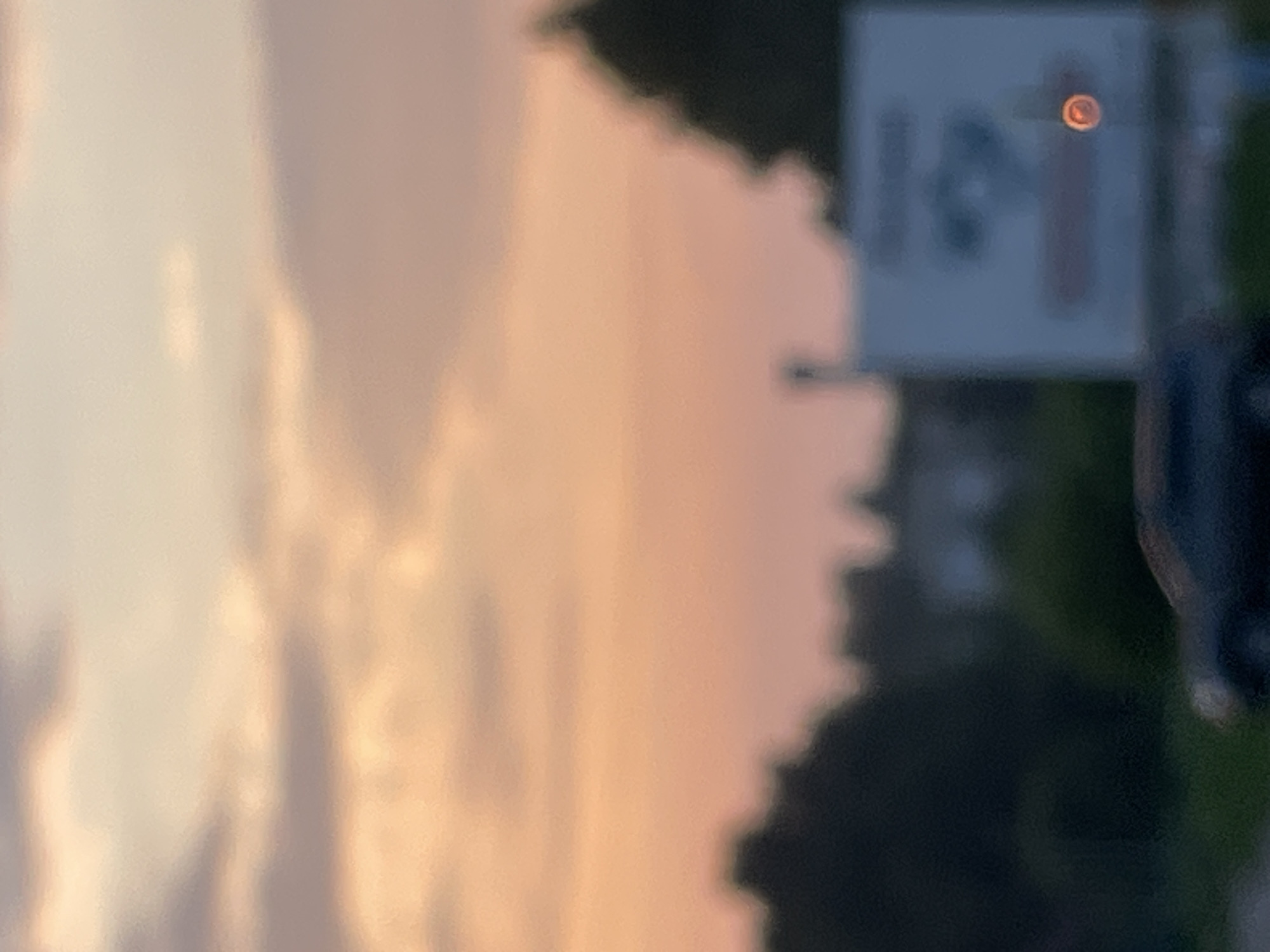 The image depicts an outdoor scene with a soft, diffused focus, characteristic of a sunset or sunrise. The sky is a gradient of pastel colors, ranging from light pinks and oranges to pale blues and grays. In the foreground, silhouetted against the sky, are dark shapes of trees and buildings. There is a white billboard with some blurry graphic elements and a small, bright orange circular light source on its right side. A dark-colored car is partially visible in the lower left portion of the image. The overall impression is of a serene, possibly rural or suburban landscape at dusk or dawn. The location is identified as Krasnye Baki, Russia.