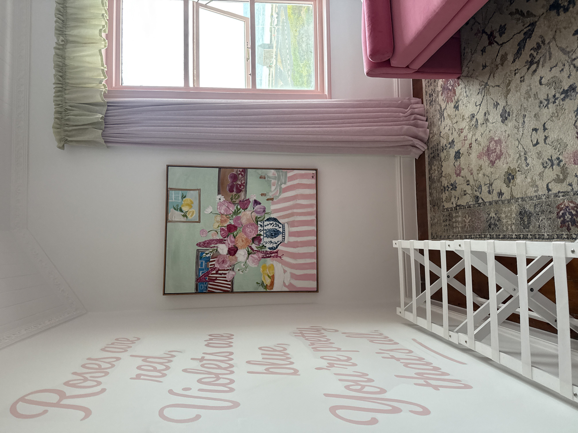 A photograph taken indoors shows a room with a window partially open, revealing a view of what could be Cape Town, South Africa, based on the coastal scenery. The room has white walls with light pink cursive writing that says, "Roses are red, Violets are blue, You're pretty that's all." Above the writing is a framed painting with floral and still-life elements, hanging beneath a window with a green ruffled valance and light purple drapes. Part of a pink sofa is visible in the upper right corner, adjacent to a patterned area rug. A white, wooden, X-frame structure stands near the lower right, angled towards the bottom of the frame.