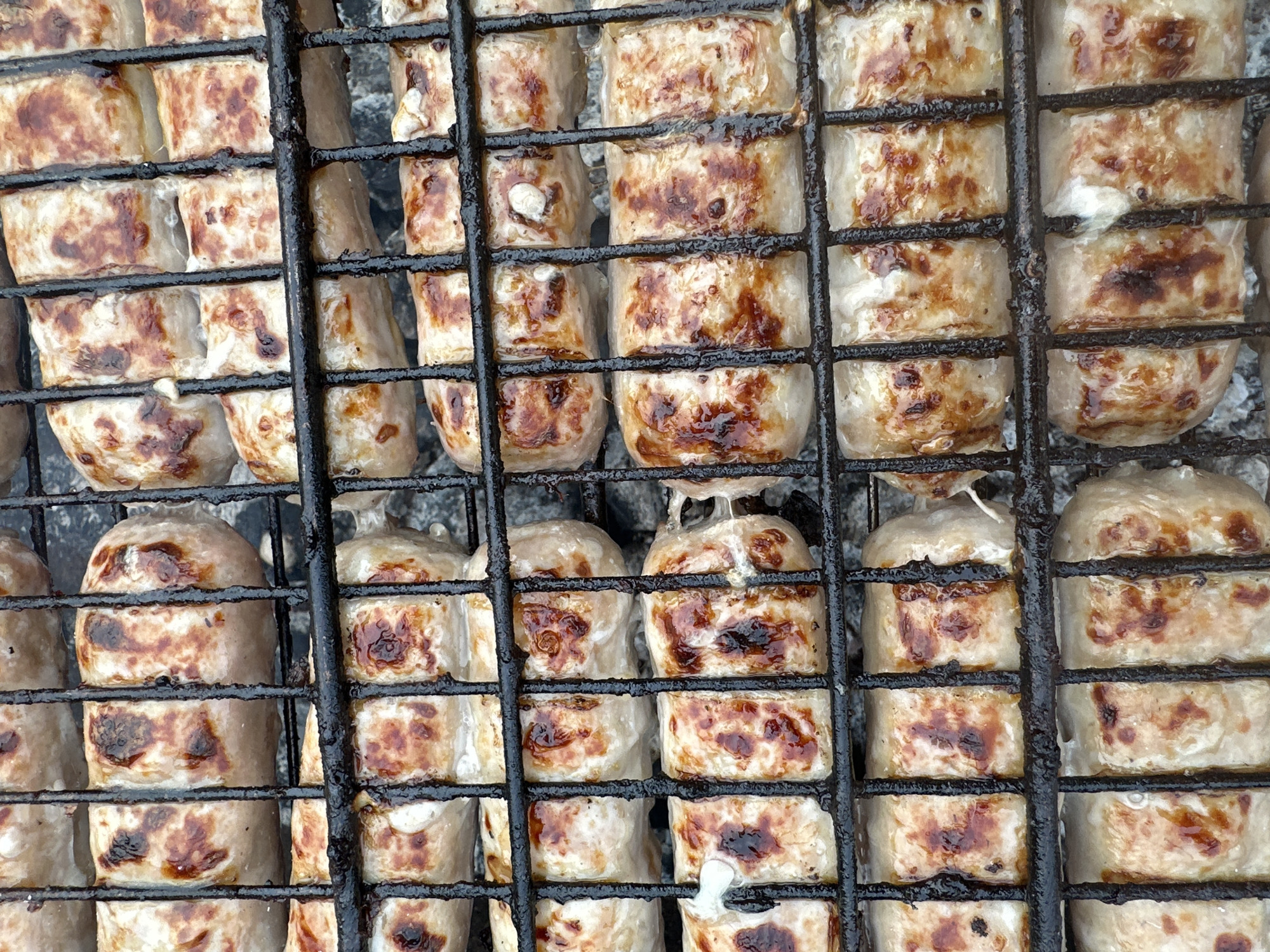 A close-up shot shows sausages on a black metal grill. The sausages are browned in places, indicating they have been cooked. The background shows faint traces of ash or charcoal, suggesting the grill is in use. The location is not mentioned and cannot be determined from the image.