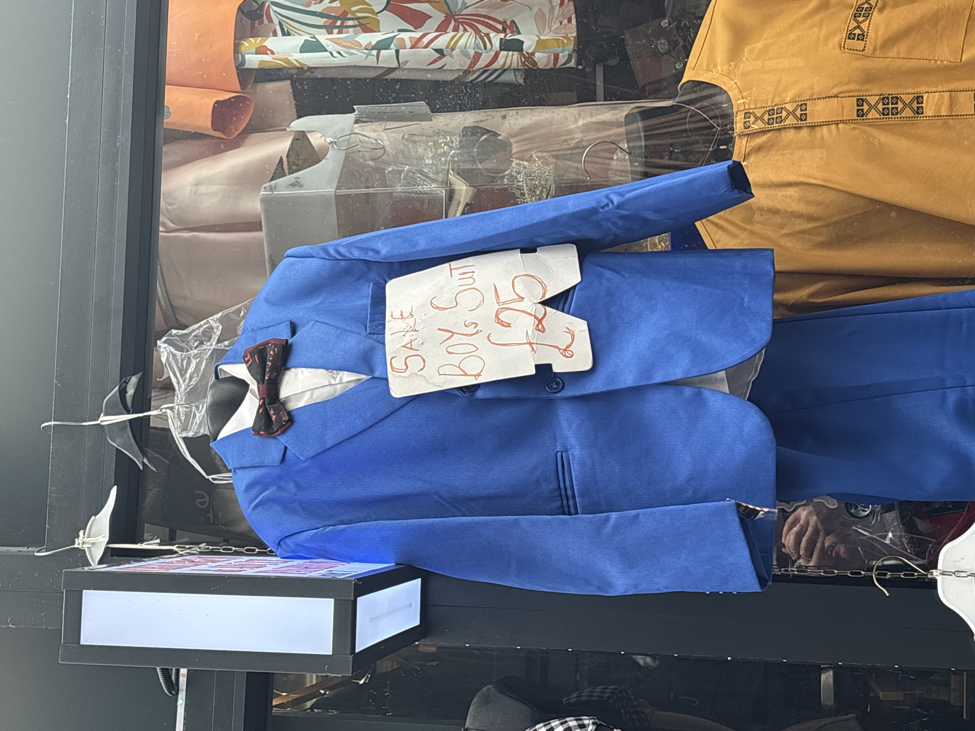 Here is a description of the image:
A medium, eye-level shot shows a blue boy's suit on sale in a shop window. A handwritten sign reading "SALE Boy Suit £75" is pinned to the suit. The suit consists of a jacket, shirt with a bowtie, and trousers, all in bright blue. The suit is displayed on a hanger.
The shop window reflects the outside environment, including some hand details and chains. Other garments are also visible in the window, including an orange/yellow top and some patterned fabrics.
The suit is placed on a black box-shaped object with a bright light strip below. The image appears to be taken in London, United Kingdom, although no explicit street indicators are shown.