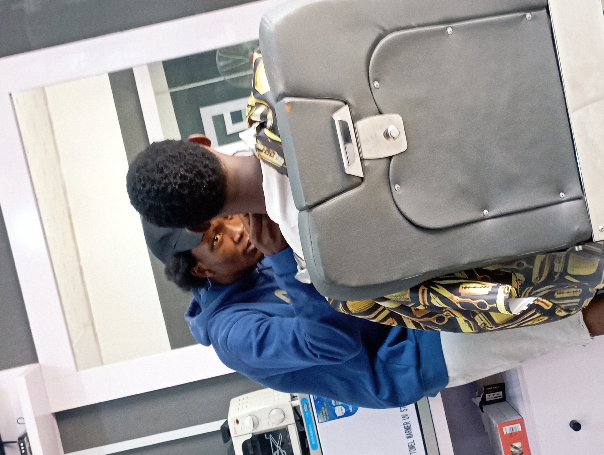 The image, which appears rotated 90 degrees clockwise from an upright orientation, captures an interior scene within a barbershop or hair salon in Jos, Nigeria.
The central subject is a young male, seen in reflection within a large, white-framed mirror. He has dark, short curly hair and is wearing a dark cap, a blue hooded sweatshirt, and a white shirt underneath. He looks forward, appearing calm and focused.
Partially visible to the right, behind the reflected subject, is another individual, presumably a barber. This person's chest and arm are visible, covered by a distinctive barber cape featuring a pattern of yellow/gold scissors, razors, and combs on a black background. The barber is positioned as if actively working on the client's hair.
The client is seated in a sturdy, dark grey barber chair, which appears to be upholstered in leather or faux leather and features metallic armrest components.
In the background, below the mirror, various salon appliances and equipment are present. To the left, a white appliance with control knobs is visible, likely a sterilizer or a similar grooming tool. To the right of that, a "TOWEL WARMER UV ST" unit is clearly discernible, further confirming the barbershop setting. The walls are predominantly light-colored, possibly white, contributing to a clean and functional environment.
