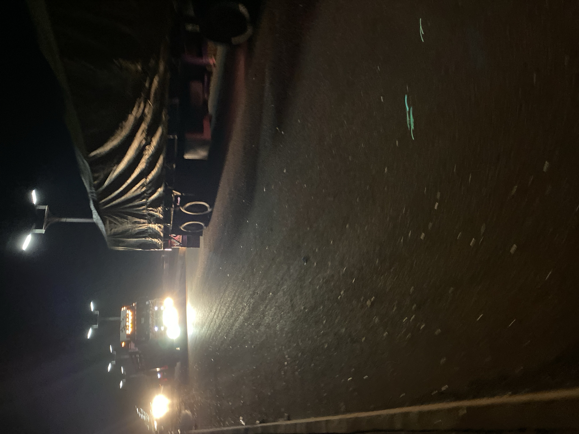 A nighttime scene showing a truck and trailer on a roadway. The location is stated to be Bukuru, Nigeria, though the image provides no locational clues. The truck's headlights illuminate the road. There are streetlights present, and the trailer of the truck is covered by a tarp.