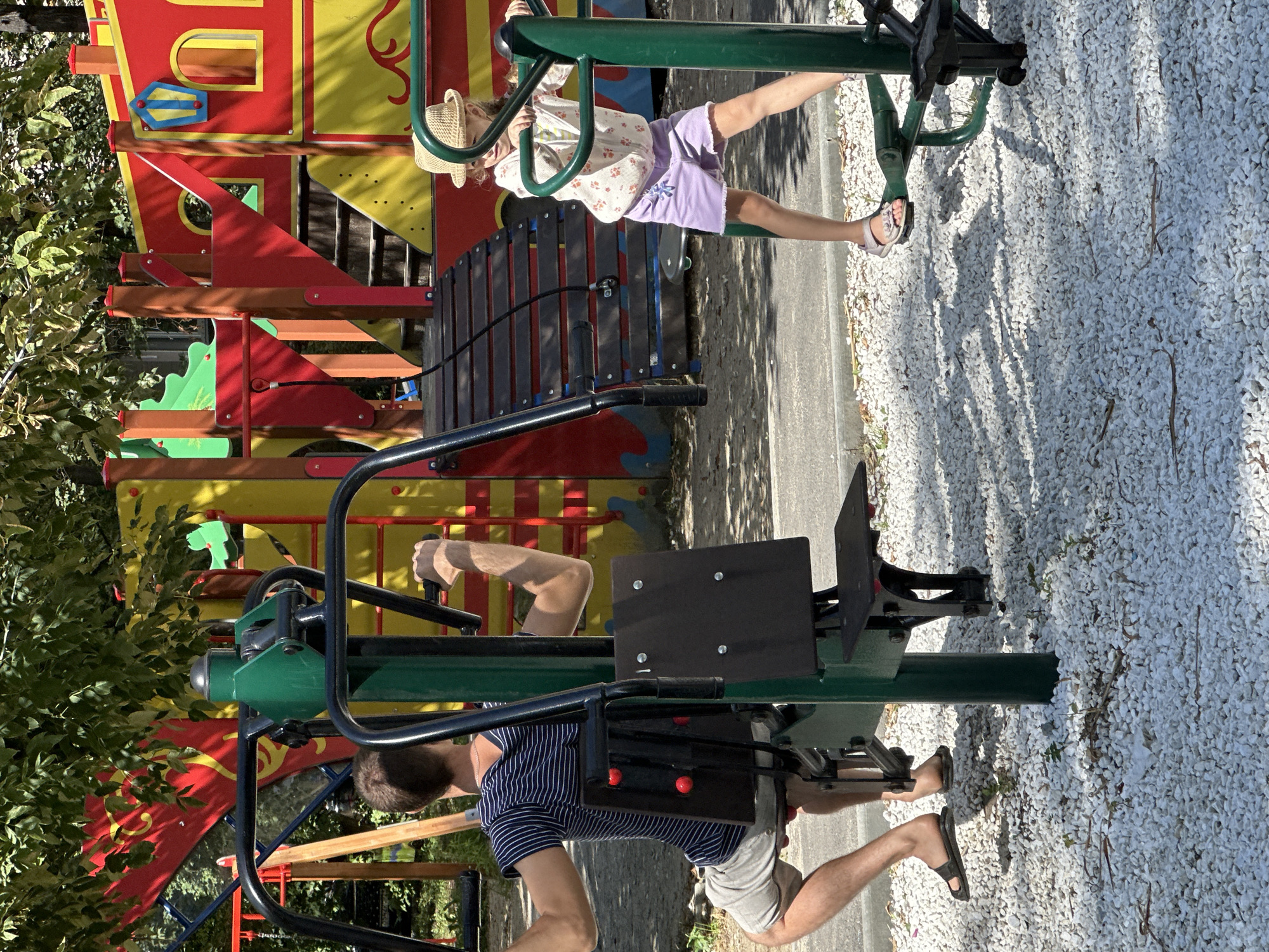 An adult male in a striped t-shirt and shorts is exercising on a green outdoor fitness machine. To his right, a young girl wearing a straw hat, a floral print top, and purple shorts is also using a similar green fitness machine. In the background, a colorful playground structure with red and yellow elements is visible. The ground is covered in white pebbles. The scene is illuminated by sunlight, casting shadows on the ground.