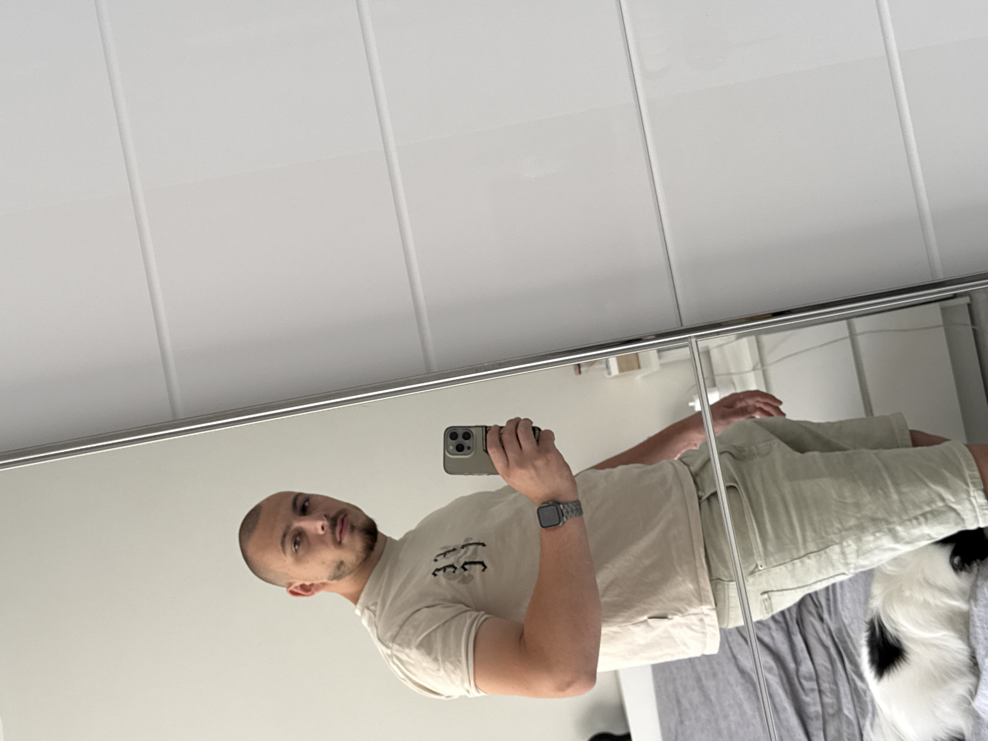 The image shows a young man with a shaved head and short beard taking a mirror selfie in what appears to be a bedroom. He is wearing a light-colored t-shirt and beige shorts. He is holding a smartphone in his right hand, and a watch is visible on his left wrist. To the left of the man, a bed with grey bedding is partially visible, and a black and white dog is lying on the bed. The background consists of a white closet with horizontal paneling. The scene is illuminated by natural light. The image does not provide any specific details or clues that would allow it to be located in Stara Zagora, Bulgaria.