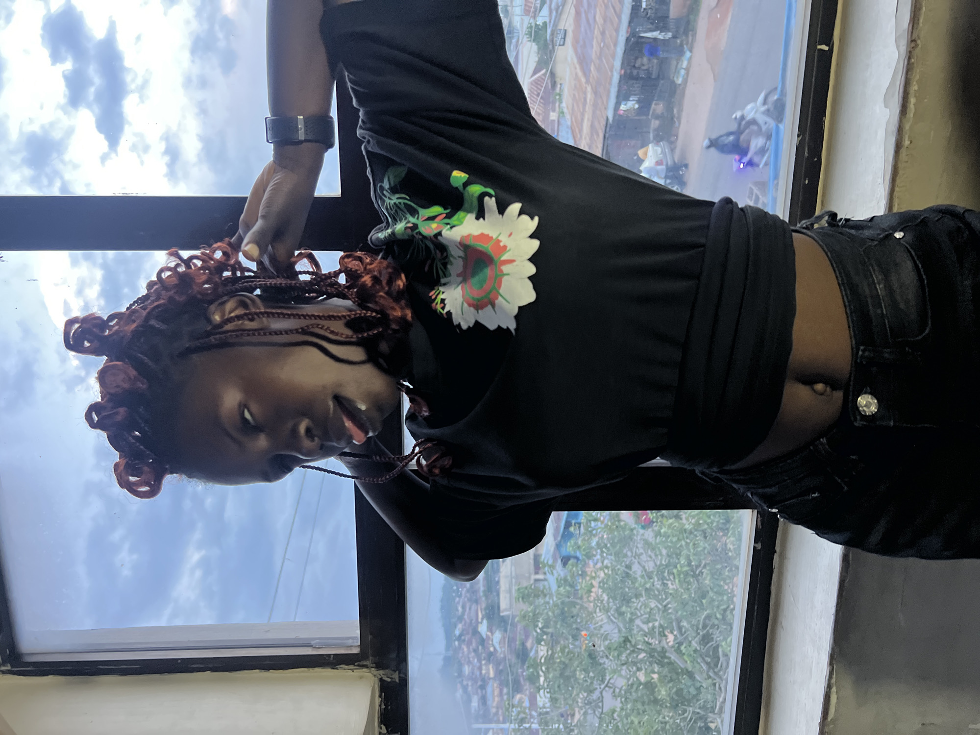 A medium shot captures a young woman with dark skin and braided hair styled in buns, posing indoors by a large window. She wears a black t-shirt with a colorful floral design and dark jeans, with her midriff exposed. Her tongue is playfully sticking out as she looks to her left, with her right hand touching her hair and left hand resting on her hip. Through the window, a cityscape is visible, characterized by red-roofed buildings, trees, and a busy street with a motorcycle and a car. The sky above is cloudy, suggesting an overcast day. The scene is likely in Jos, Nigeria, given the architectural style and urban landscape.