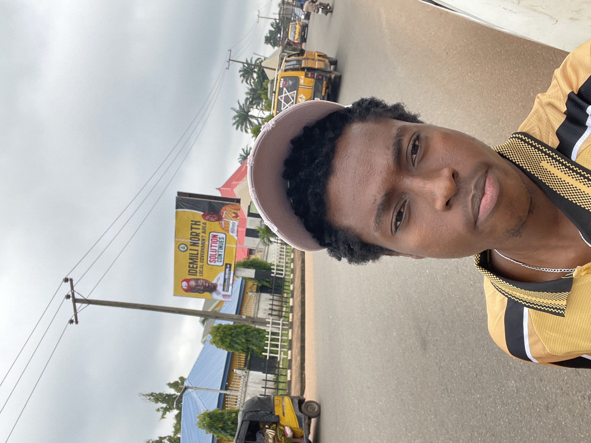 The image is a selfie taken outdoors, with the camera tilted to capture the subject and the surrounding environment. The subject is a young Black man wearing a light-colored baseball cap and a yellow and black patterned shirt. He is looking towards the camera with a neutral expression.
In the background, the scene depicts a street in Nkpor, Nigeria. Several yellow minibuses, commonly used as public transport, are visible on the road, along with a yellow and black auto-rickshaw. A large billboard featuring a smiling man in a red cap and the text "IDEMILI NORTH LOCAL GOVERNMENT AREA" and "SOLUTION CONTINUES" is prominently displayed. Power lines stretch across the sky, which is overcast, suggesting a cloudy or possibly rainy day. Palm trees and some greenery are scattered throughout the scene. The overall impression is of a bustling urban environment in Nigeria.