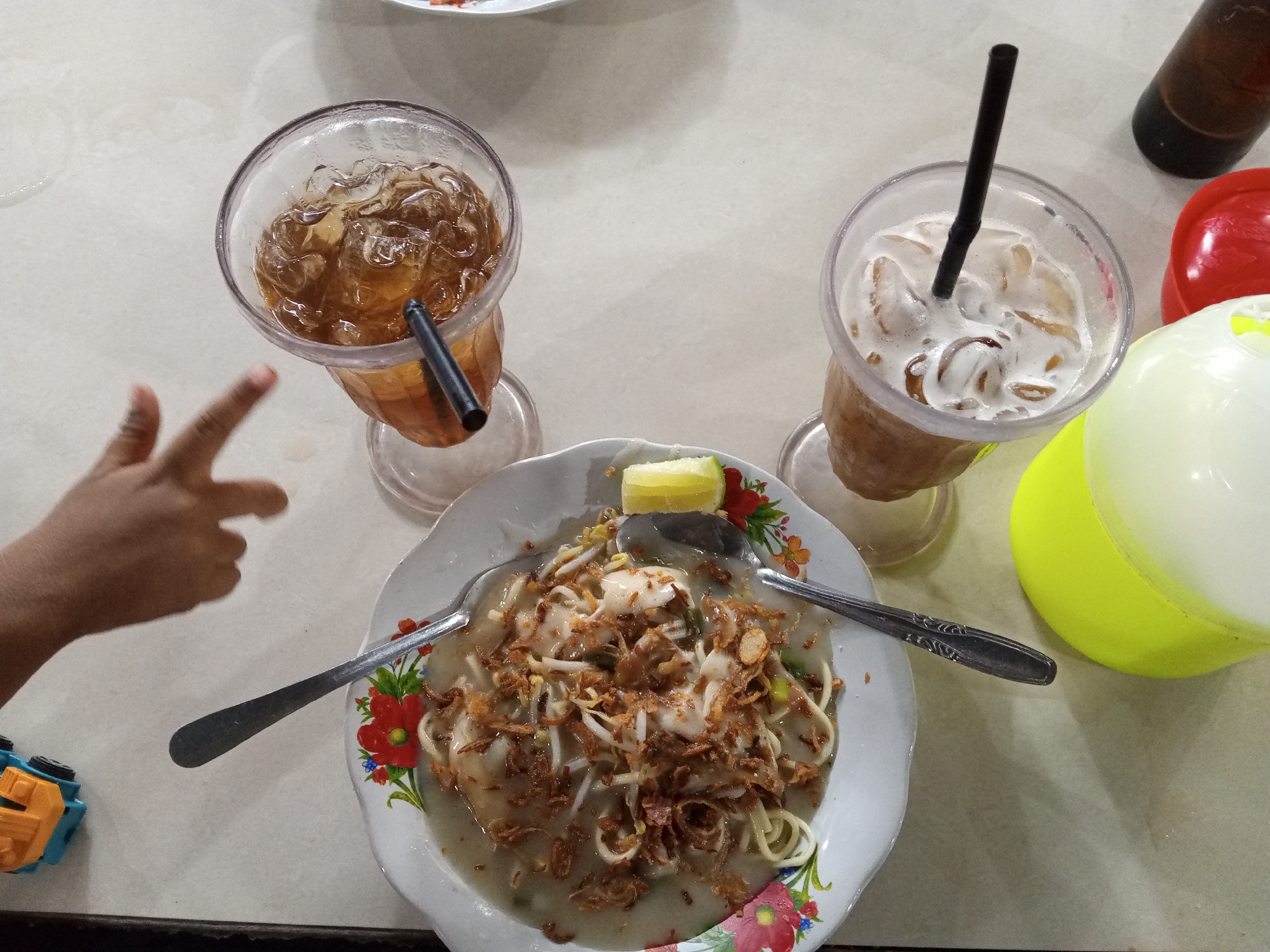 Two glasses of iced tea, one with ice and a black straw, the other with foam, ice, and a black straw, sit on a white table. A bowl of noodles with bean sprouts and fried onions, a lemon wedge, and two spoons are in the center of the table. A child's hand is to the left of the bowl, and a blue and yellow toy truck is to the left of the hand. A red lid and a yellow and white container are to the right of the bowl. The scene appears to be a restaurant in Gampongteungoh, Indonesia.