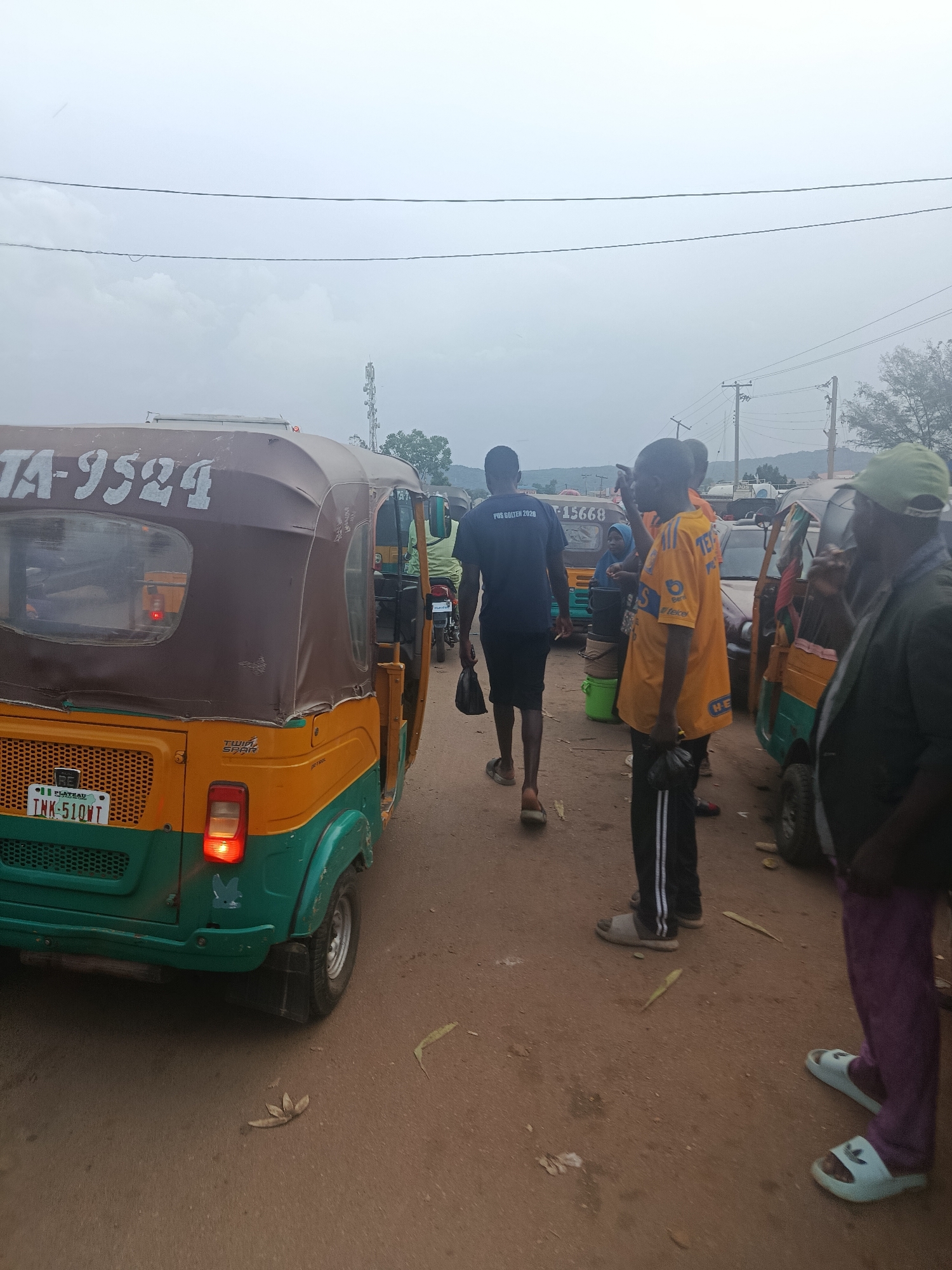 The image depicts an outdoor scene, possibly in Rafin Par, Nigeria, with a cloudy sky overhead and power lines crossing the frame. The ground is a reddish-brown dirt or gravel. In the foreground, the rear of a yellow and green three-wheeled vehicle, commonly known as an auto-rickshaw or Keke Napep, is visible. It has the license plate "INK-51QWT" and "TA-9524" is written on the top.
To the left of the vehicle, a person wearing a dark blue t-shirt with white text that reads "PDS GOLTEN 2020" and black shorts is walking away from the camera. They are carrying a black bag in their left hand and wearing sandals.
To the right of the auto-rickshaw, a group of people are standing around. One individual is wearing a bright yellow t-shirt with "TE" and other text in darker colors, black pants with white stripes, and is carrying a black bag. Another person in the group is wearing a green cap and a dark jacket. A woman wearing a light blue headscarf and a dark colored garment is also present, holding a green bucket. Further to the right, a person in purple pants and white slides is standing.
In the background, more auto-rickshaws and a few other vehicles are present. A cell tower is visible in the distance, along with some trees and hills. The overall impression is of a busy street or transportation hub.