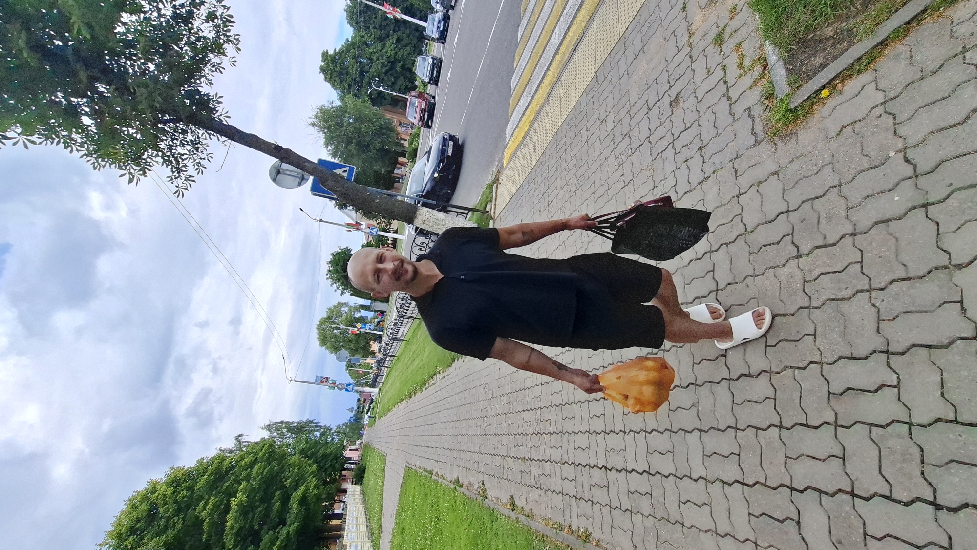 A bald, smiling Caucasian man in a black polo shirt and black shorts walks on a paved sidewalk. He is carrying an orange plastic bag filled with items in his left hand and a dark tote bag in his right hand. He is wearing white slide sandals. The sidewalk is made of interlocking concrete pavers and is lined with grass on the left and a street on the right. Traffic can be seen on the street, and there are streetlights and signs visible. A large tree with green leaves extends over the sidewalk from the right. In the background, there are more trees, buildings, and a cloudy sky. The location appears to be a street in Myadzyel, Belarus, evidenced by the characteristic architecture and street signage.