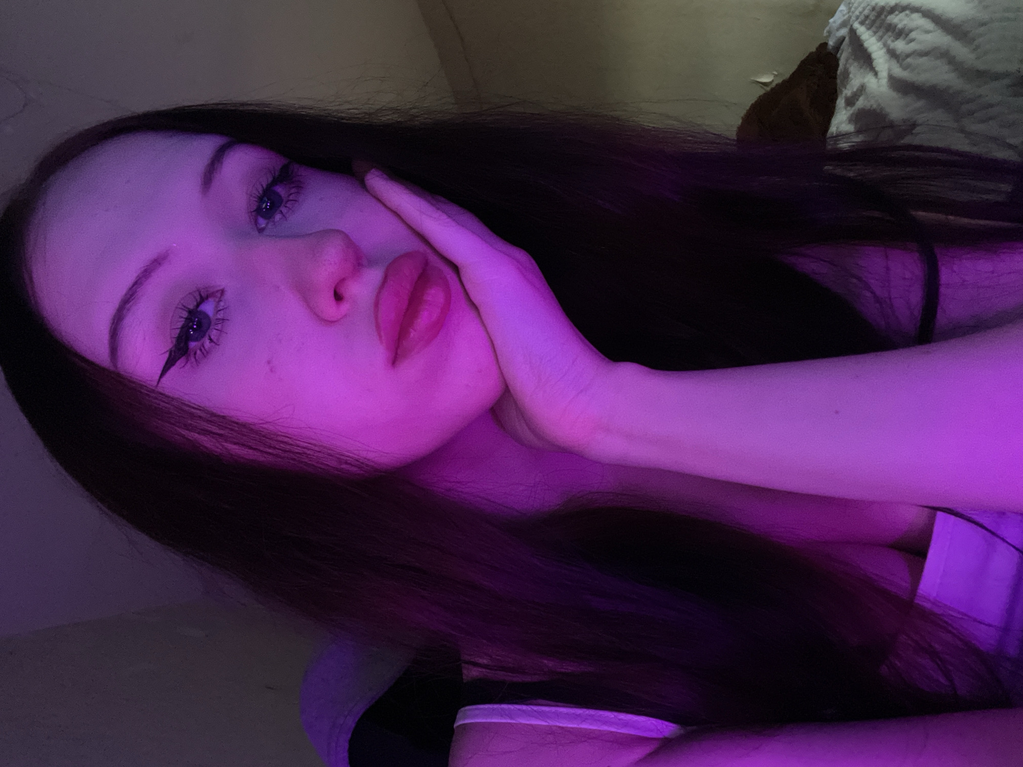 A young woman with long, dark hair is shown in a close-up shot, illuminated by purple lighting. She is resting her cheek on her hand, with her eyes looking slightly upward and her lips parted. She is wearing a white top with thin straps. The background appears to be a dimly lit room, possibly a bedroom, with a textured wall and a white, possibly quilted, surface in the lower right corner, which might be bedding. The scene is likely captured indoors, possibly in Votkinsk, Russia, given the context.