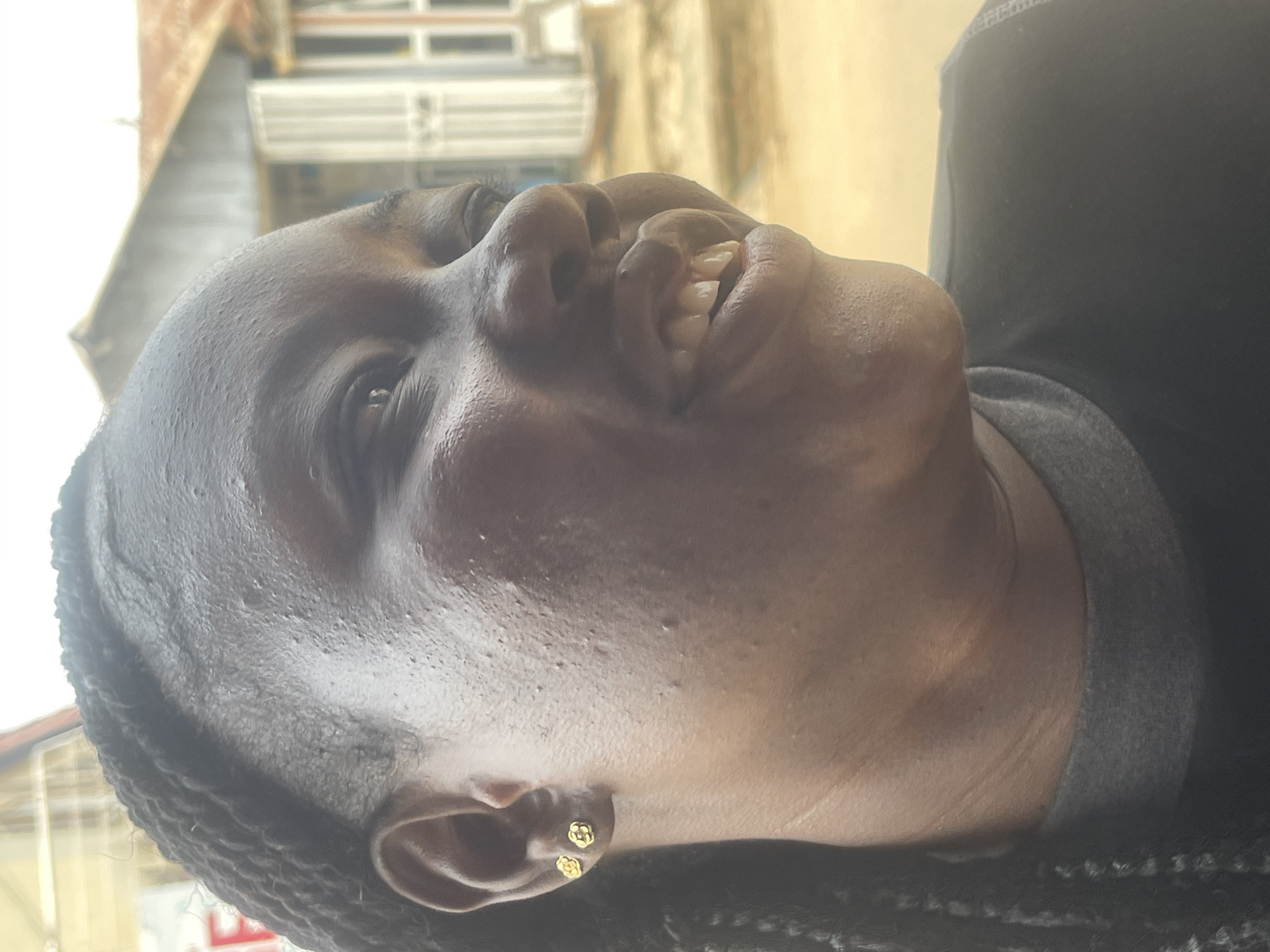 A medium close-up shot shows the right side of a Black woman's face, who is wearing a gray and black t-shirt and two small gold flower-shaped stud earrings in her right ear. Her braided hair is visible along the top and back of her head. Her skin appears slightly oily and has some minor texture. The woman is smiling, revealing her teeth. The background is slightly blurred, showing a building with a corrugated metal roof and white doors. The scene appears to be outdoors in an urban environment, possibly in Abuja, Nigeria.