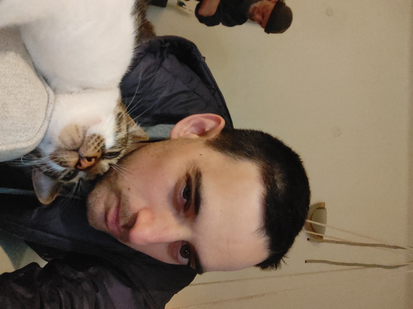 A man is lying down with a cat resting on his chest. The cat is white and tabby, with its head turned sideways. The man has short dark hair and is looking at the camera. The background is a plain wall. A blurry image of another person is visible at the top of the frame. The location and city are unknown.