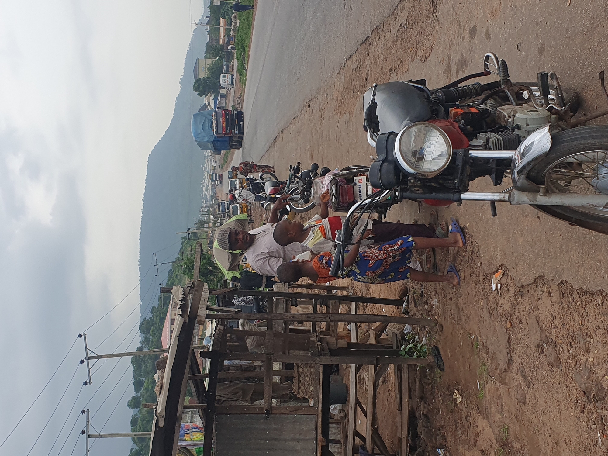 The image captures a roadside scene in Lokoja, Nigeria, under a cloudy sky. In the foreground, a red and black motorcycle is parked on a dirt and asphalt shoulder. To the left, a makeshift wooden stall with corrugated metal siding displays various goods, including what appear to be stacked cardboard items. A man in a white shirt and a checkered cap stands near the stall, talking to two children. One child, a boy, wears a striped shirt and dark pants. The other child, a girl, is dressed in a colorful patterned dress and blue flip-flops. Behind them, a line of parked motorcycles and a few other individuals are visible. The road stretches into the distance, with traffic including trucks, vans, and smaller vehicles. The background features lush green hills and scattered buildings, indicating an urban or semi-urban environment. Power lines with insulators are strung across the scene, supported by wooden poles. The ground is a mixture of reddish-brown dirt, gravel, and patches of asphalt.